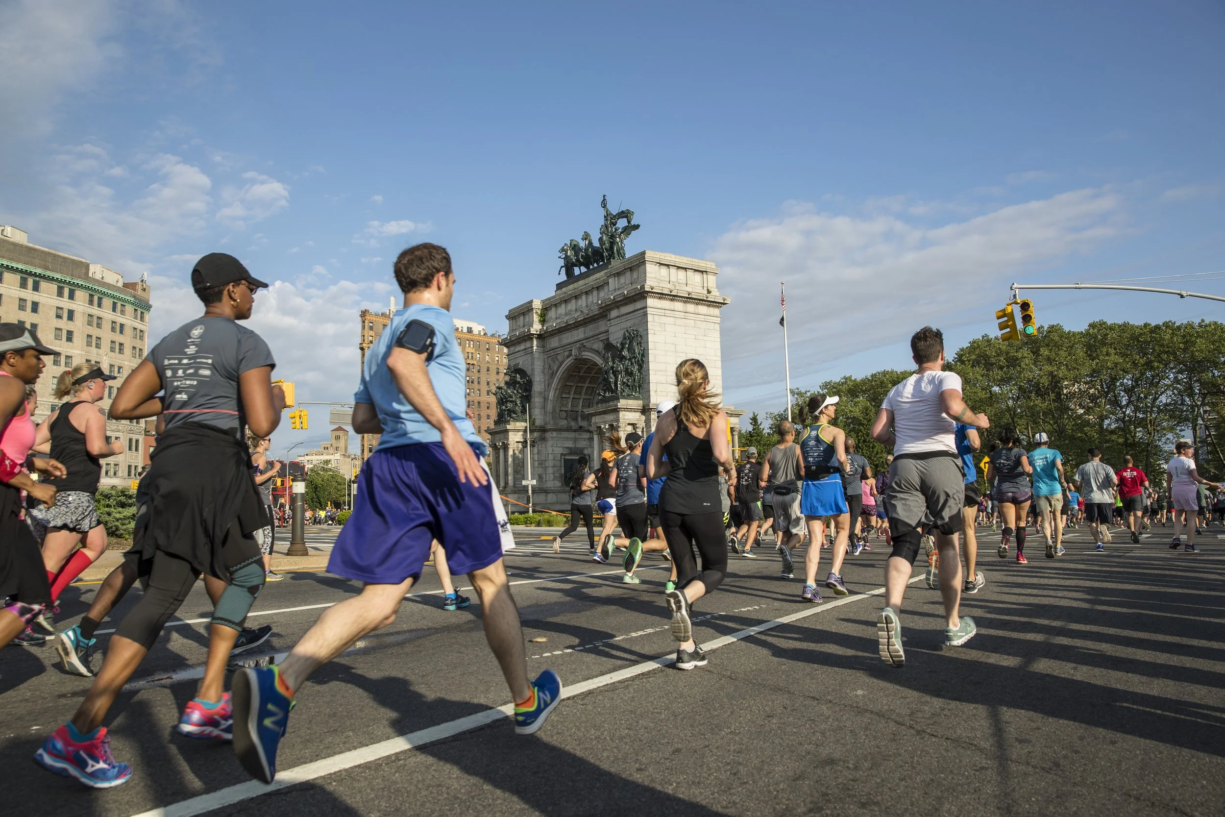 A group of people running in a marathon or race event on city streets with historic buildings and a monument in the background, under a blue sky with scattered clouds.