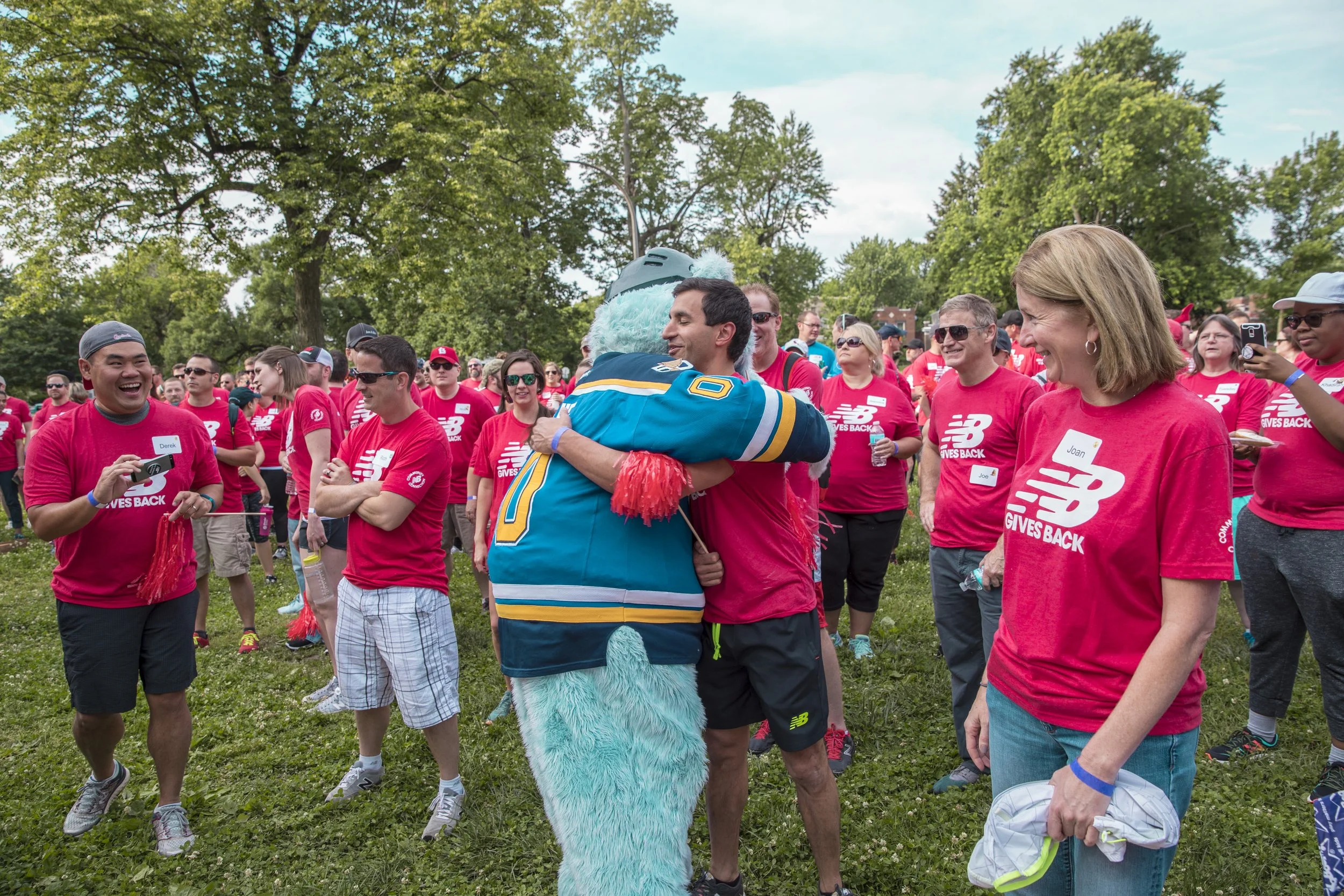 A group of people wearing red shirts with 'GIVES BACK' written on them gathered outdoors in a park with trees. A person in a blue furry mascot costume is hugging a man.