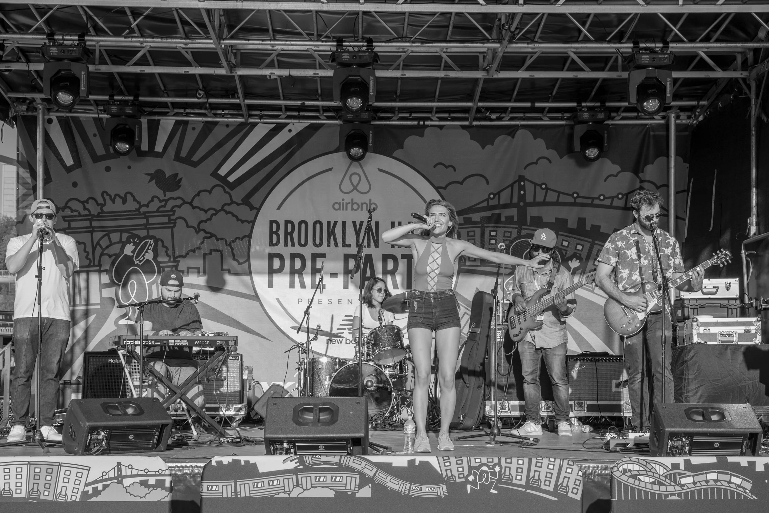 A band performs on stage at an outdoor event in Brooklyn, with a backdrop advertising the Brooklyn Pre-Party by Airbnb. The band consists of a singer, a woman with a microphone and guitar, a drummer, a bass guitarist, and a keyboard player. The stage is equipped with sound equipment and lighting.