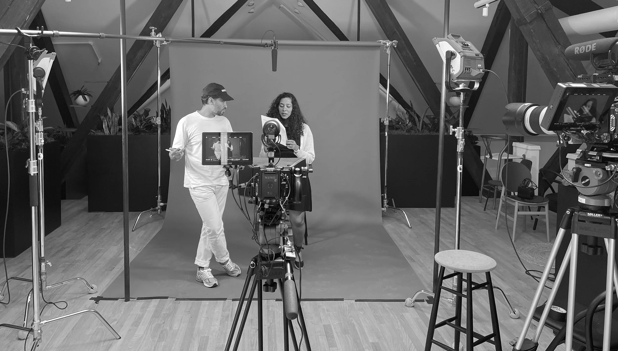 Two people standing in front of a gray backdrop in a video production studio, engaged in a conversation or reading, surrounded by professional filming equipment including cameras, lights, and a boom microphone.