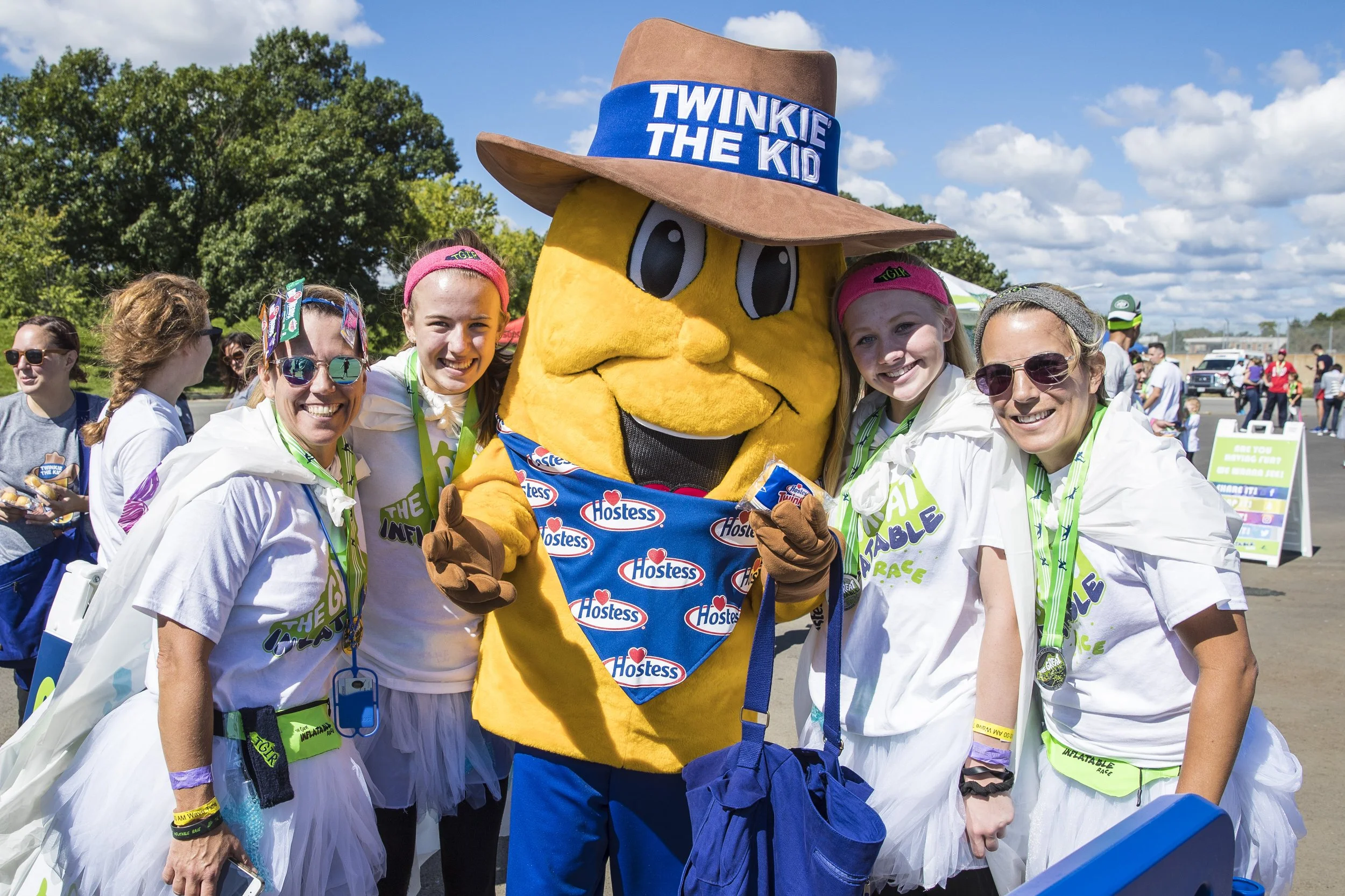 A group of women and a mascot dressed as Twinkie the Kid at an outdoor event. The mascot is wearing a large cowboy hat with a blue band that says 'Twinkie the Kid' and a costume covered in Hostess logo stickers. The women are smiling, some wearing sunglasses and event shirts, standing together with a clear blue sky and some trees in the background.