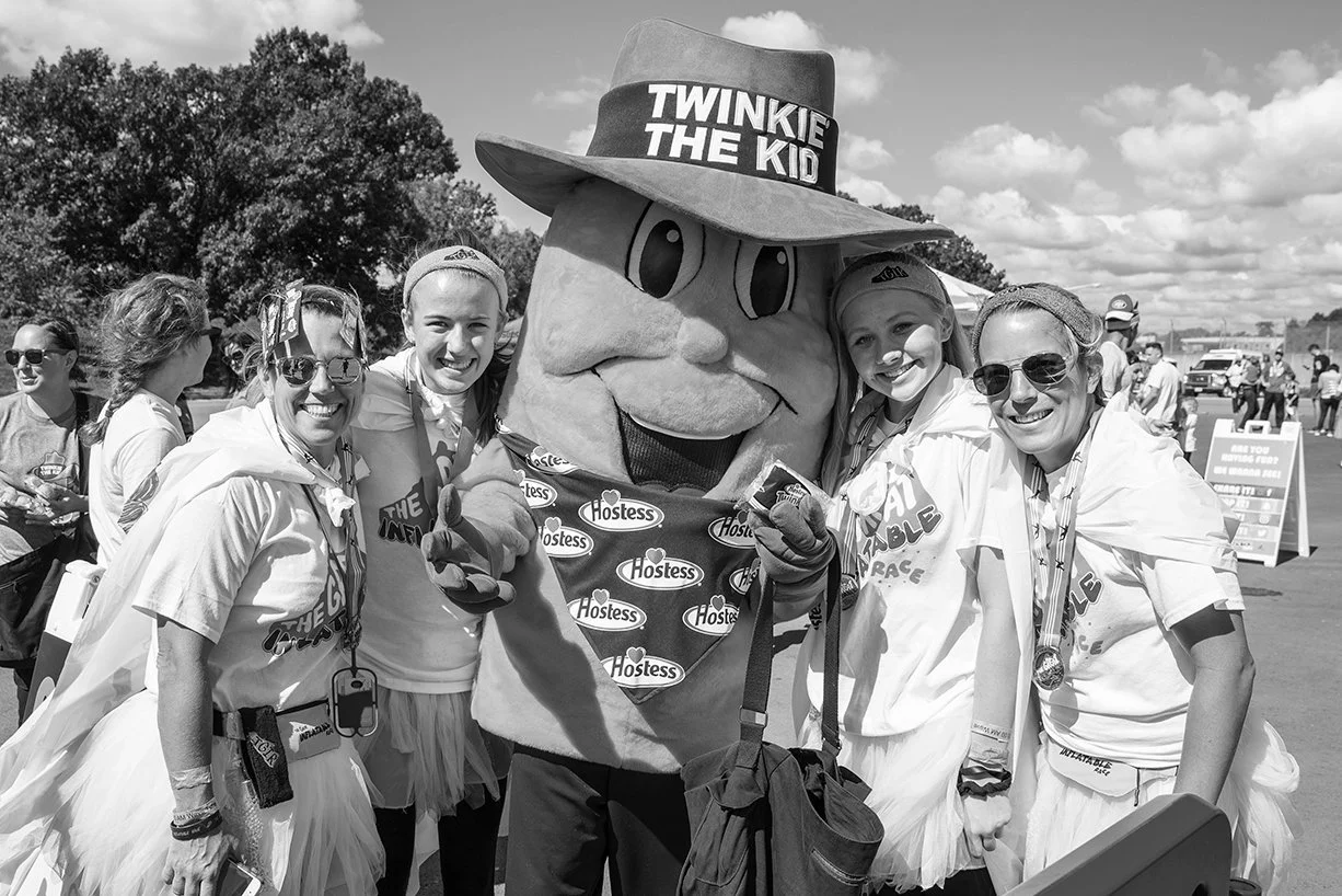 Women and girls wearing race t-shirts and medals, standing with the mascot of Twinkie the Kid, a giant Twinkie snack cake with a cowboy hat that says 'Twinkie the Kid' and a bandana that says 'Hostess', at an outdoor event.