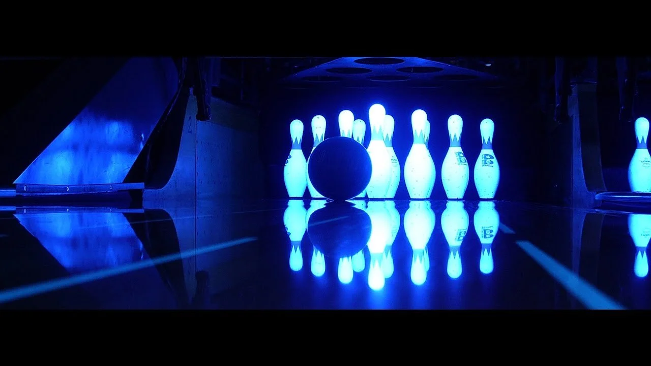 A bowling ball approaching glowing blue bowling pins on a dark lane.
