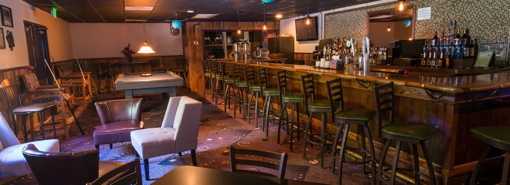 Empty bar with wooden counter, high chairs, and a few chairs and tables in a lounge area with dim lighting.