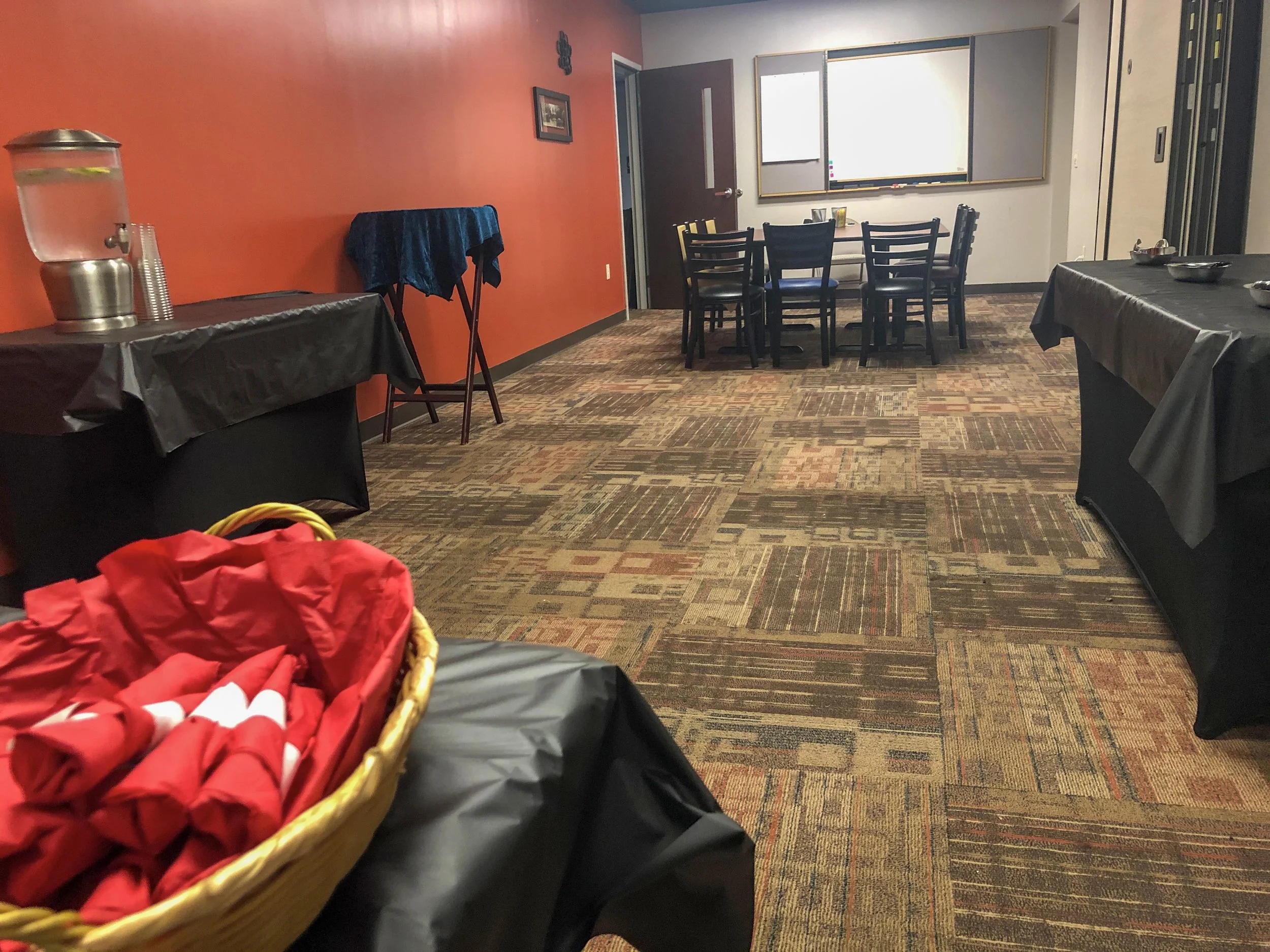 Empty conference room with tables covered in black tablecloths, a water dispenser, a basket with red cloth, a table with drinks, a whiteboard, and chairs arranged around a table.