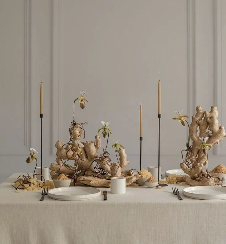 Elegant dining table with large ginger root centerpieces, surrounded by four place settings with plates, forks, and small bowls, tall candles in black holders, and minimalist decor in a neutral-colored room.