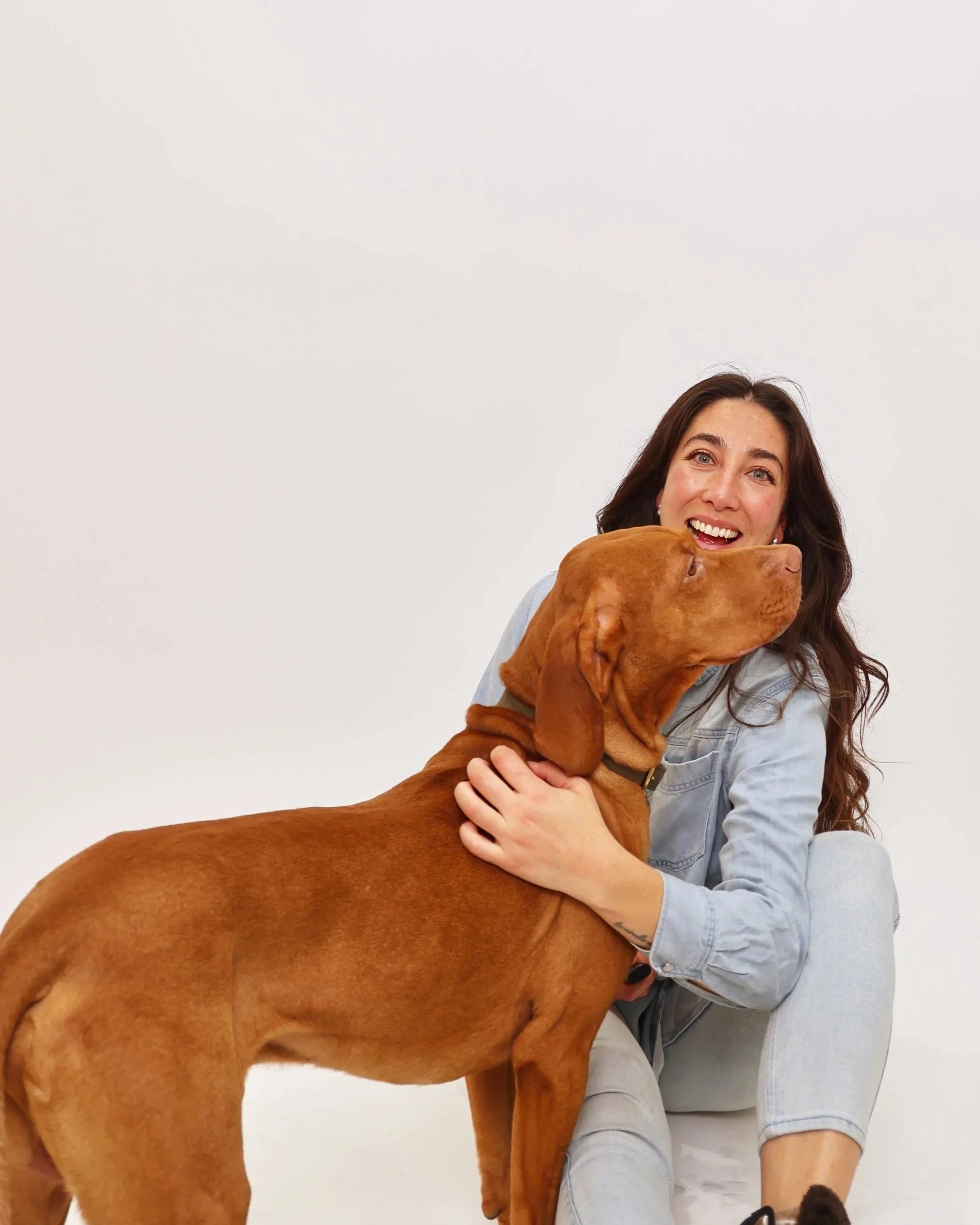 A woman with long, dark hair, wearing a light denim jacket and gray shorts, is hugging a large brown dog while sitting on the floor against a plain light-colored wall. The woman is smiling with visible teeth.