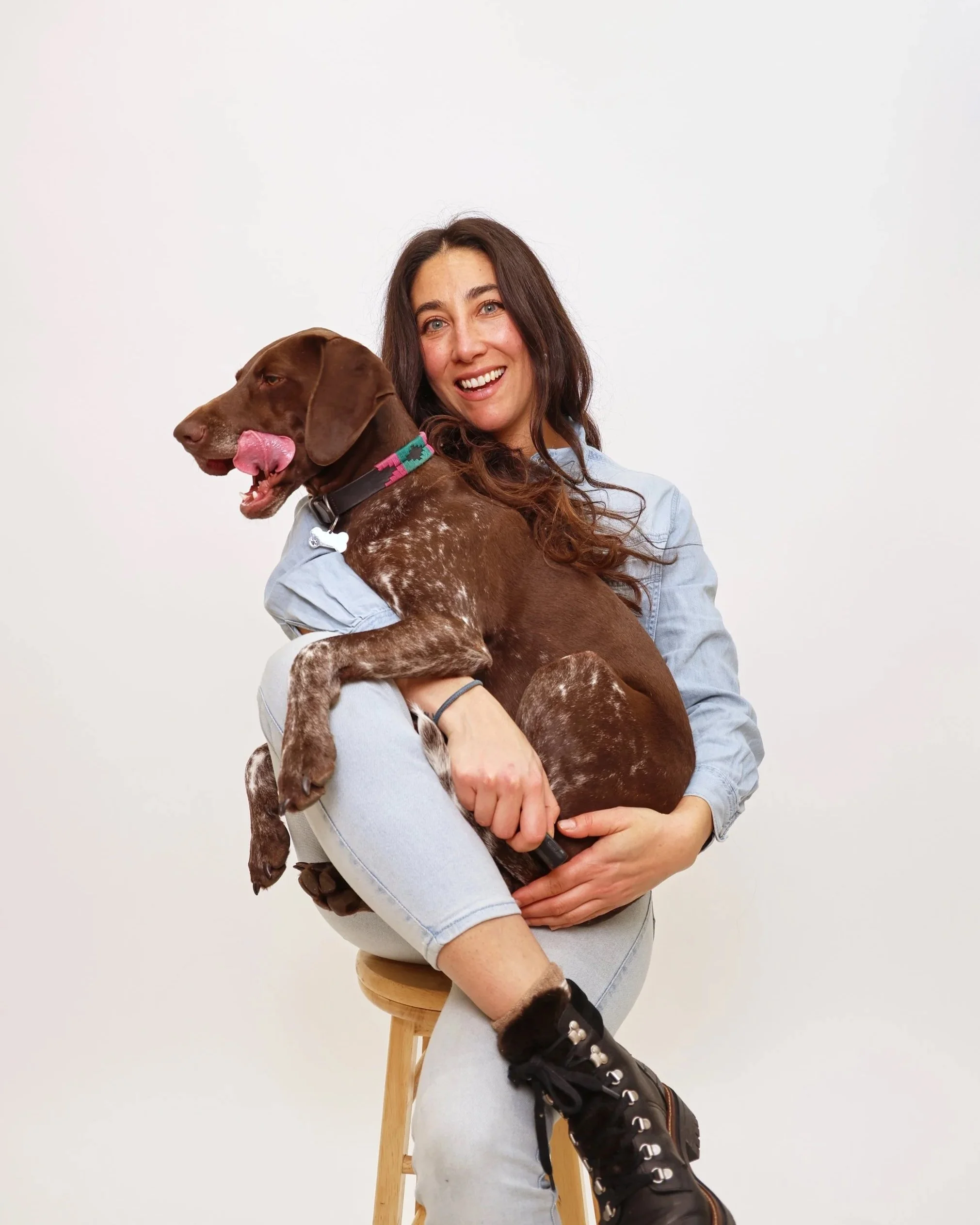 A woman with long dark hair, wearing a light blue shirt, is sitting on a wooden stool holding a large brown and white dog. The dog has a pink and green collar and is licking its nose. The background is plain white.