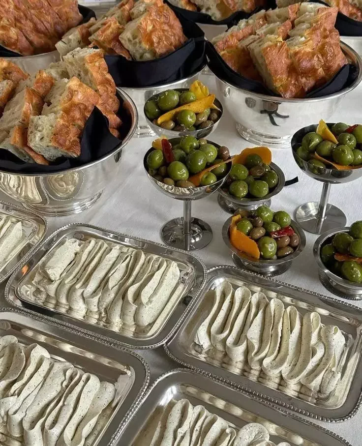 Tray of piped white mousse or frosting, bowls of green olives and pickled vegetables, and bowls of sliced bread or pastries.
