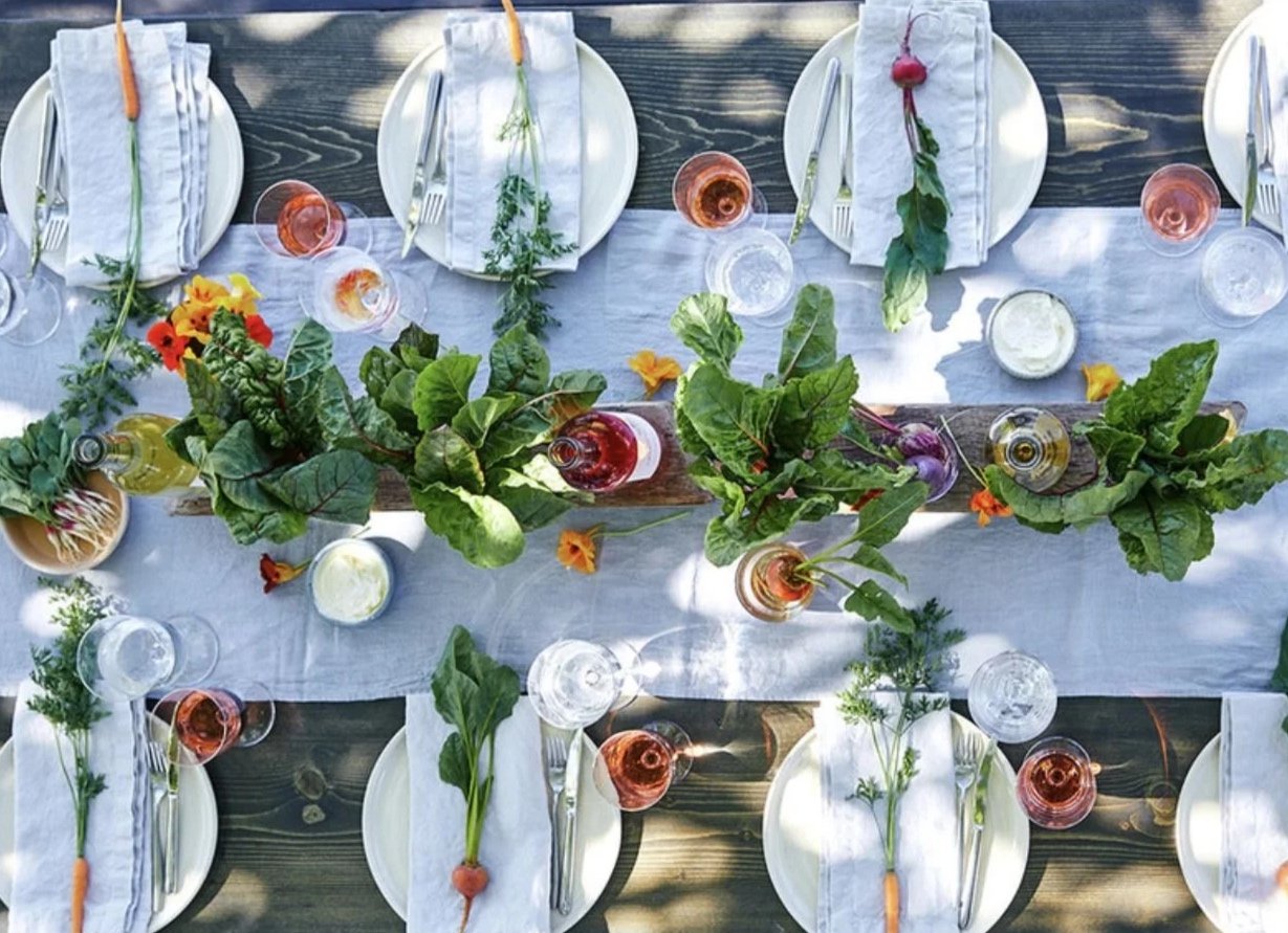 Overhead view of a table set for a meal, decorated with fresh greens and flowers, with plates, glasses, and utensils arranged neatly.