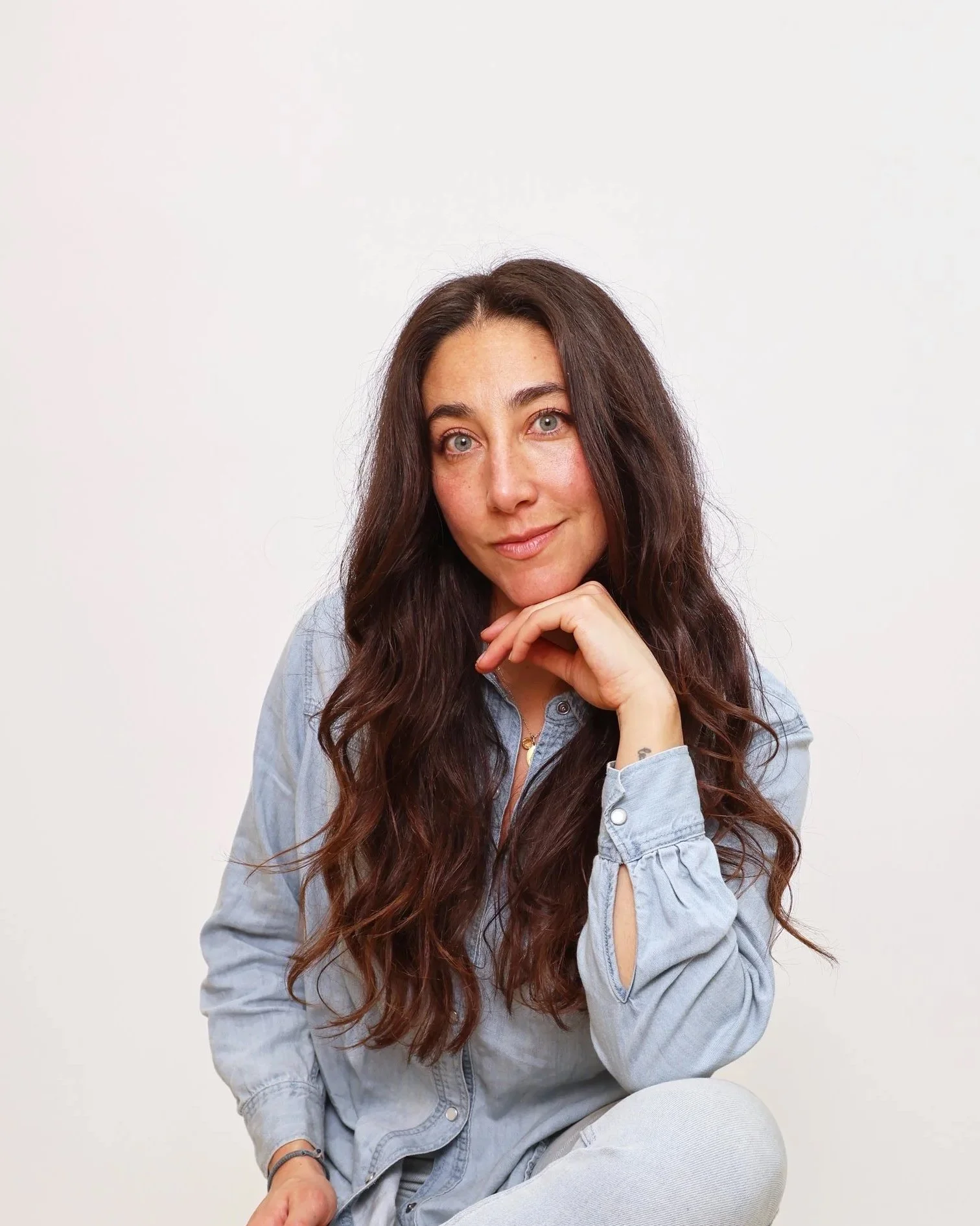 A woman with long, wavy dark hair and blue eyes, wearing a light denim shirt, sitting against a plain white background, resting her chin on her hand and smiling slightly.
