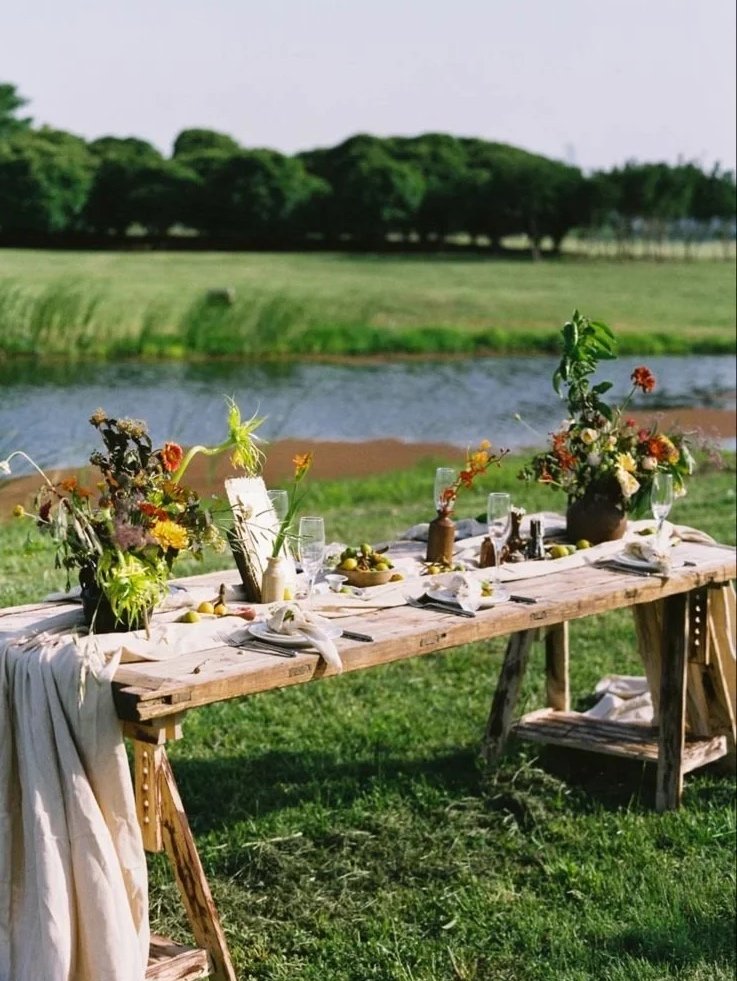 A rustic outdoor dining table set on grass near a body of water, decorated with colorful flower arrangements in vases, plates, glasses, and napkins.