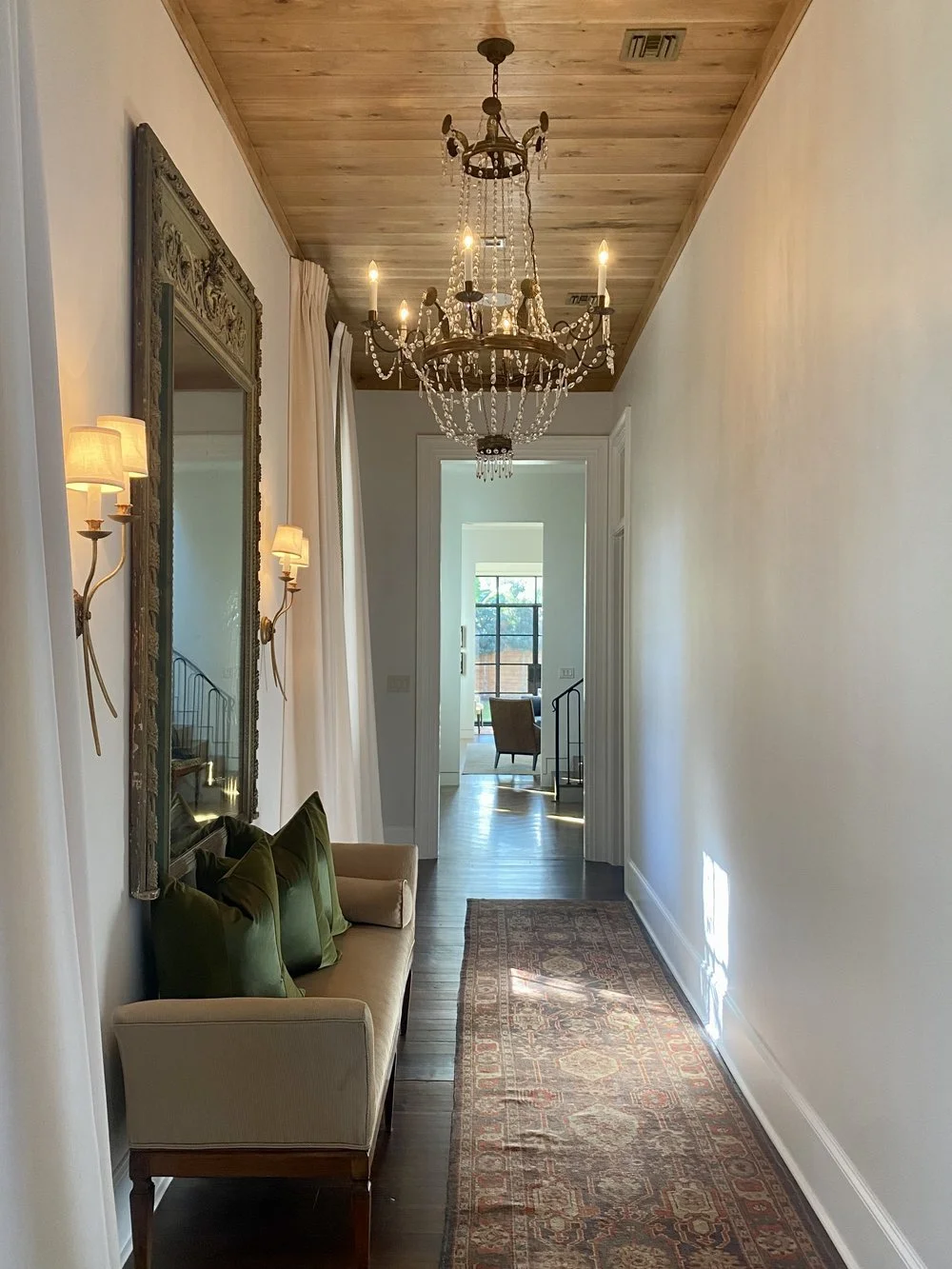 A hallway with wooden flooring, a chandelier hanging from a wooden ceiling, wall sconces beside a mirror, a beige sofa with green and beige pillows, and a long patterned rug, leading into a room with large windows and a chair.