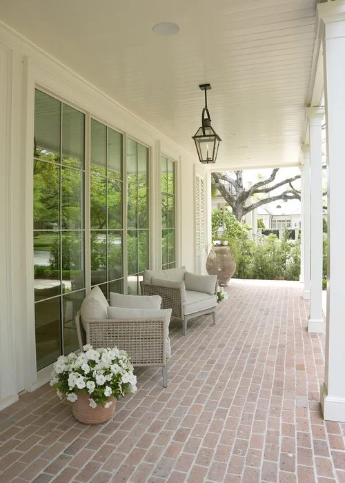 Covered porch with white wicker furniture, large potted plant, hanging lantern, and a view of trees and houses outside.