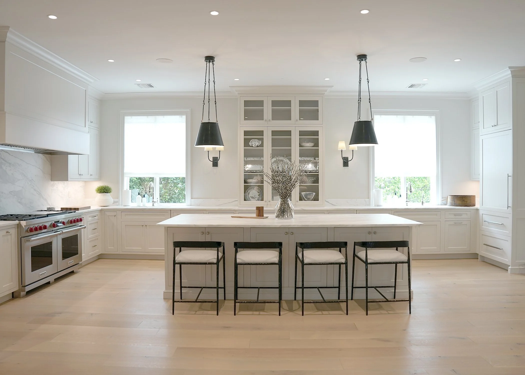Bright modern kitchen with white cabinetry, a large island with seating, black pendant lights, and two windows with white blinds.