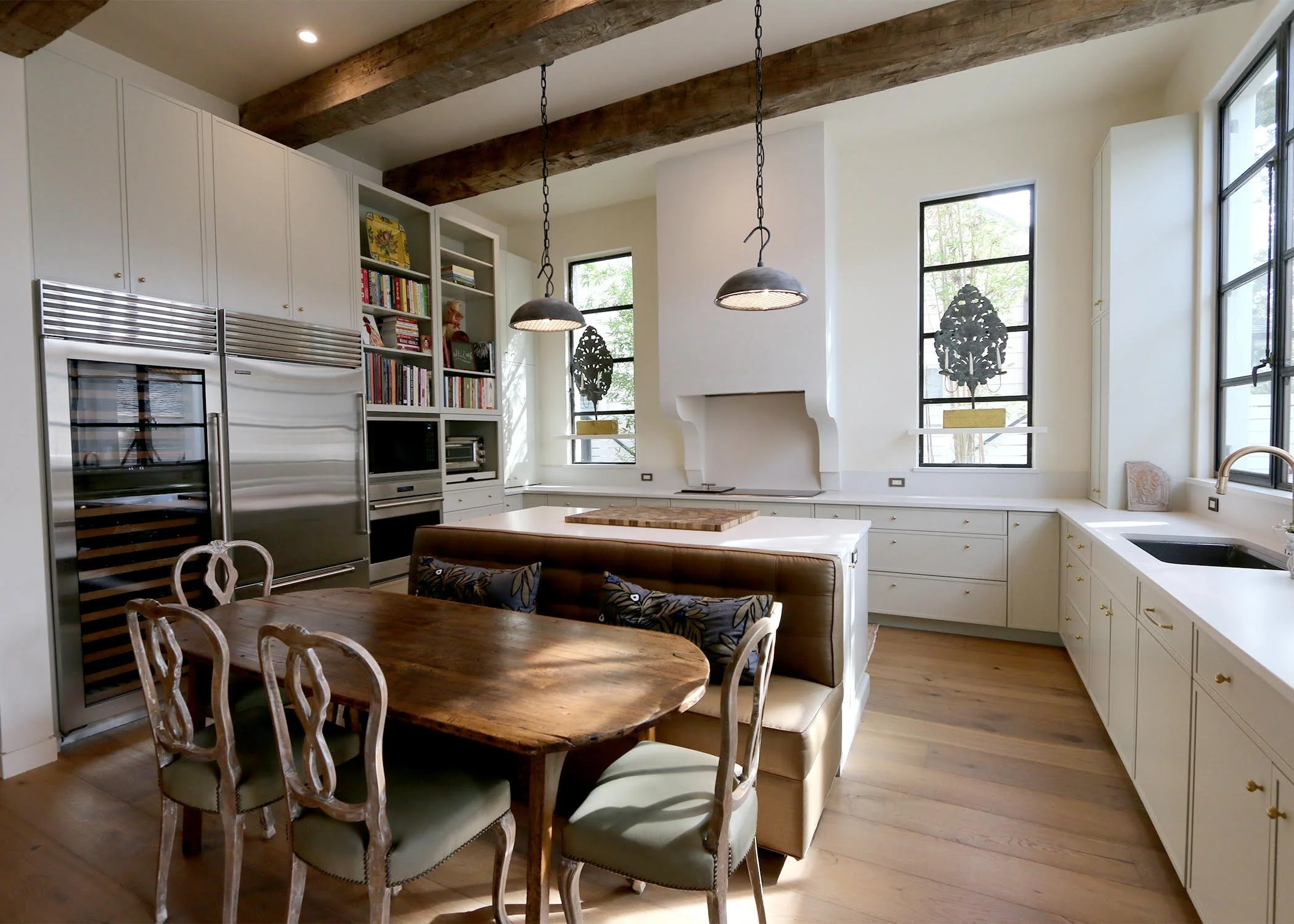 Modern kitchen with white cabinets, wooden beams on the ceiling, and large windows with black frames. There is a wooden dining table with chairs and a built-in bench. Two pendant lights hang above the table, and a fireplace with decorative black scro