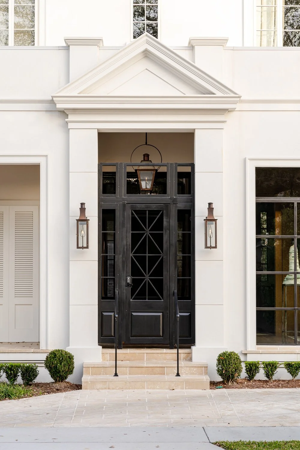 Front entry of a white house with a black front door, flanked by wall-mounted lantern lights, small shrubs, and a set of steps leading up to the door.