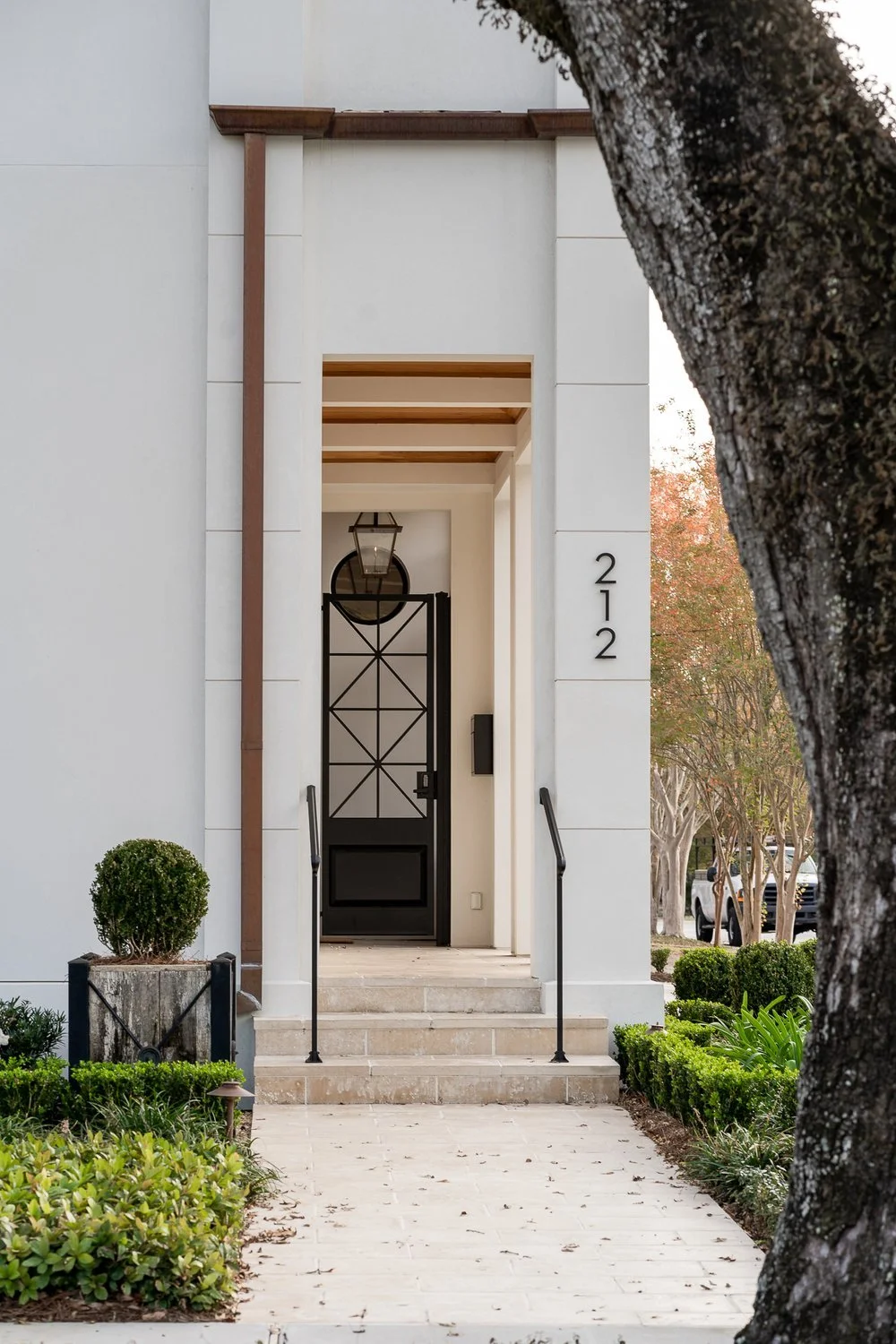 Modern house entrance with steps, black gate, and house number 212, surrounded by greenery and trees.