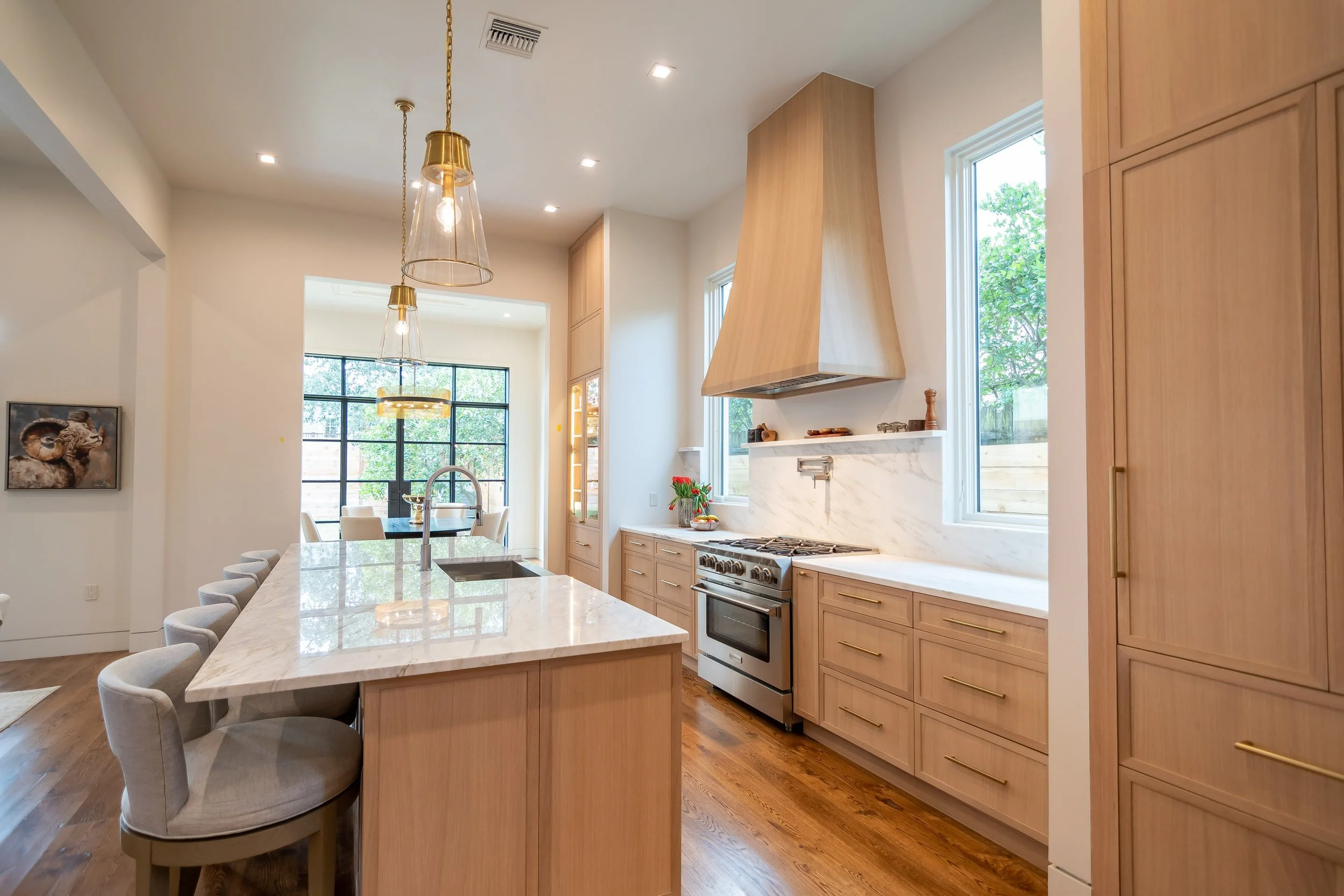 Modern kitchen with wooden cabinets, white marble countertops, a stainless steel stove, and pendant lights over a kitchen island with beige upholstered chairs.