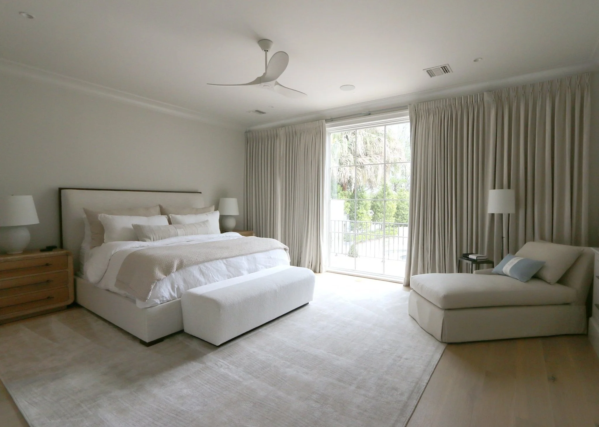 Bright bedroom with a large bed, white bedding, wooden nightstands, two lamps, side chaise lounge with cushion, large window with cream curtains, view of trees outside, ceiling fan, light-colored carpet and hardwood floors.