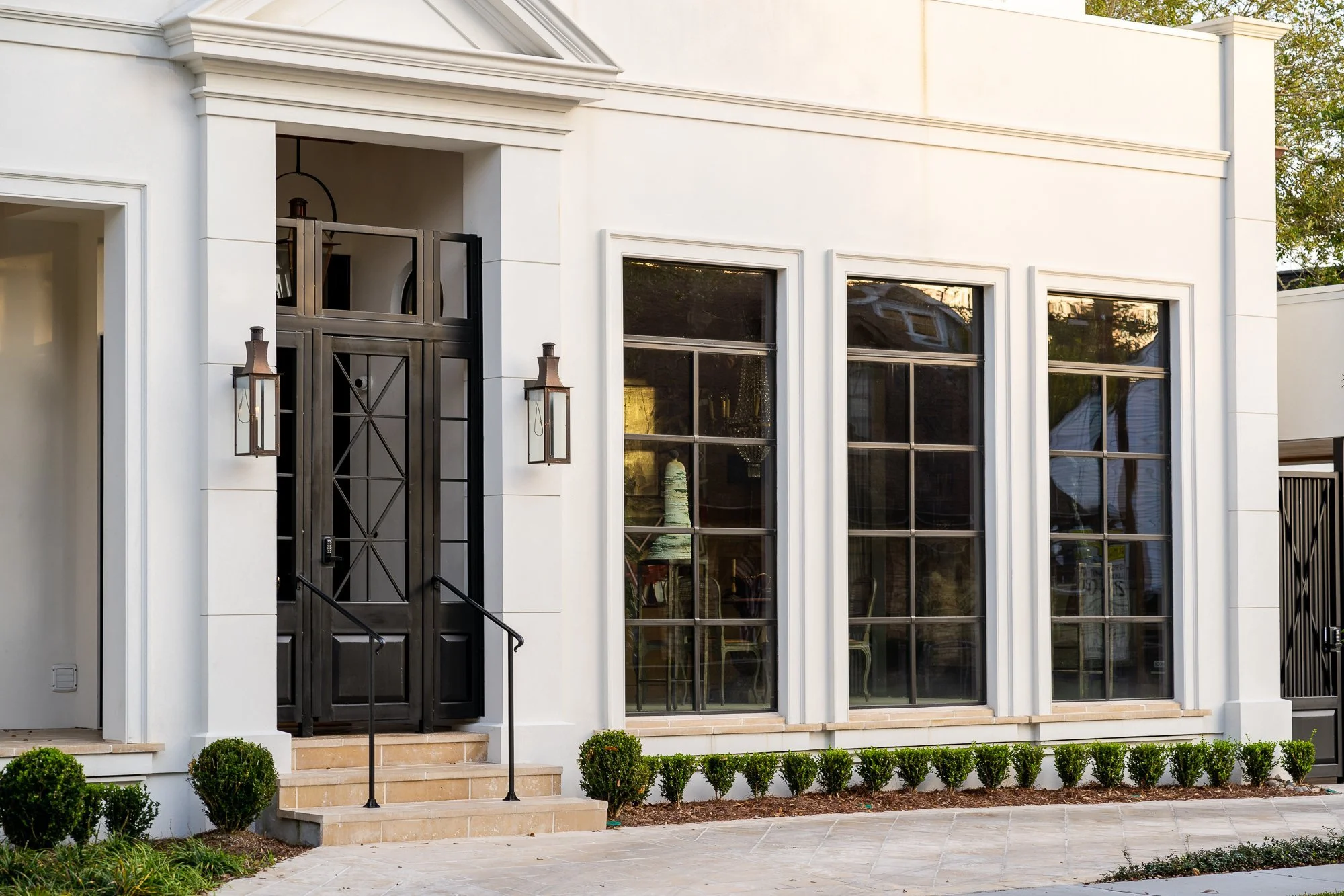 Front exterior of a modern white house with three tall windows, a black security door with steps, and landscaped bushes.