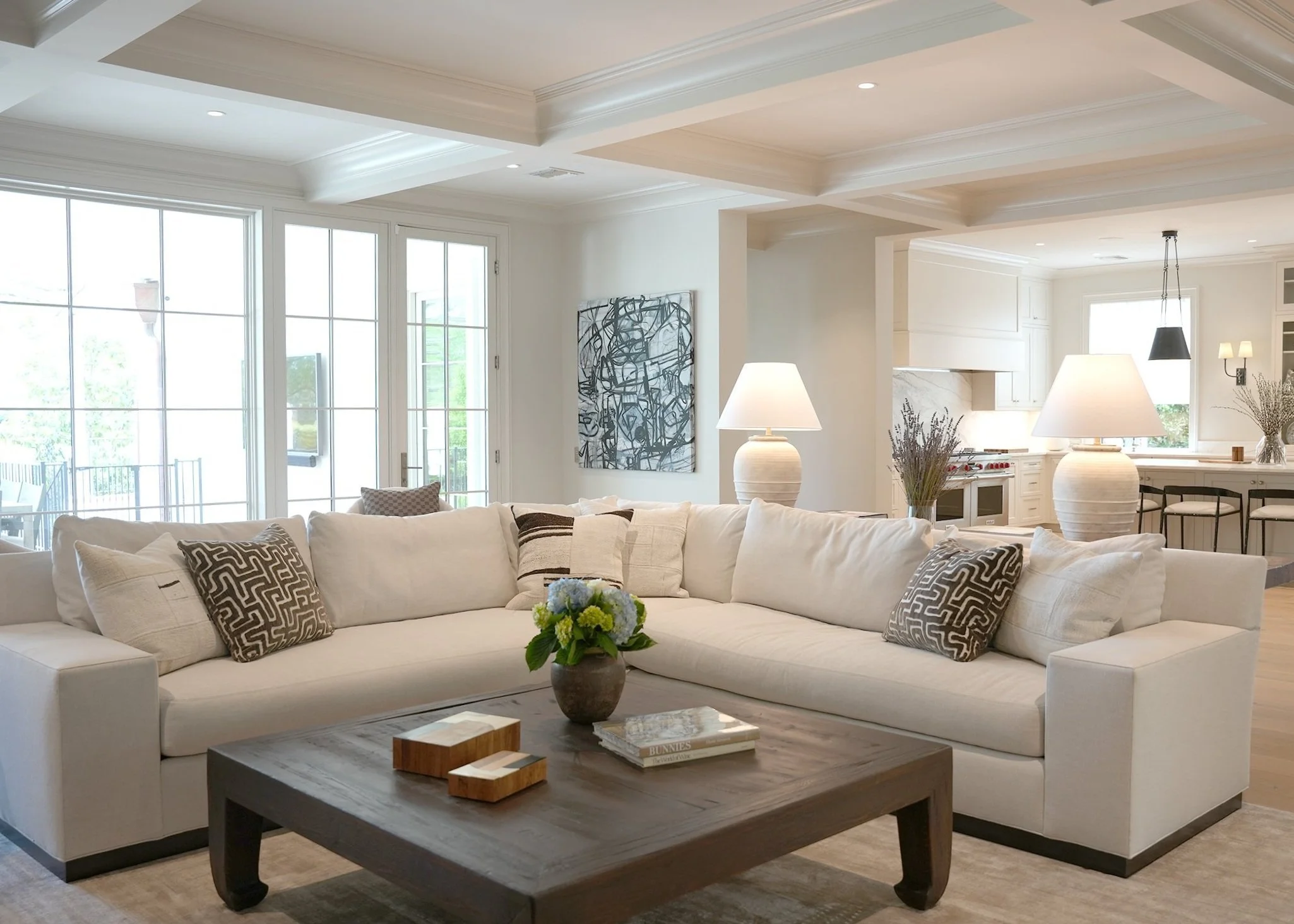 Bright, airy living room with a large white sofa adorned with patterned and solid throw pillows, a wooden coffee table with decorative items, and a kitchen in the background with white cabinetry, pendant lights, and a dining area.