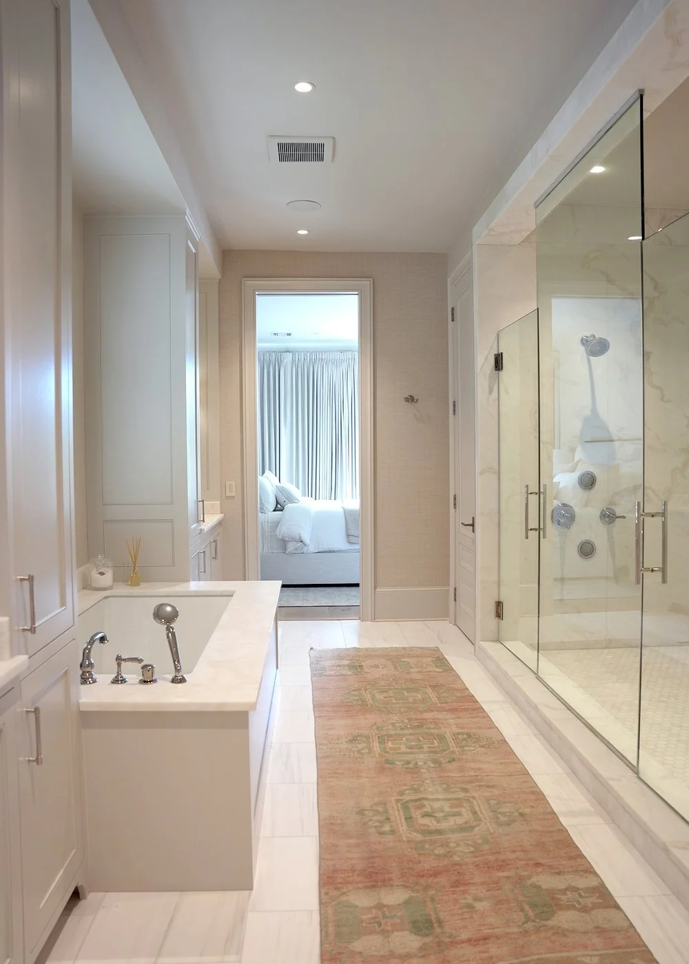 Bathroom with a marble shower enclosed by glass doors, a bathtub, and a view into a bedroom with a white bed and curtains.
