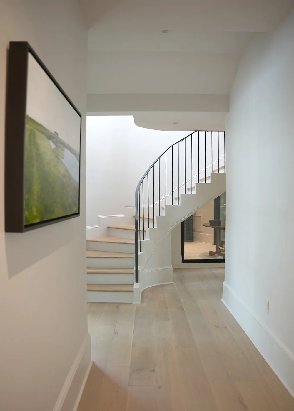Interior view of a modern home with a curved staircase featuring white risers and black railing, light wooden flooring, a framed picture on the wall, and a room visible through a glass window.