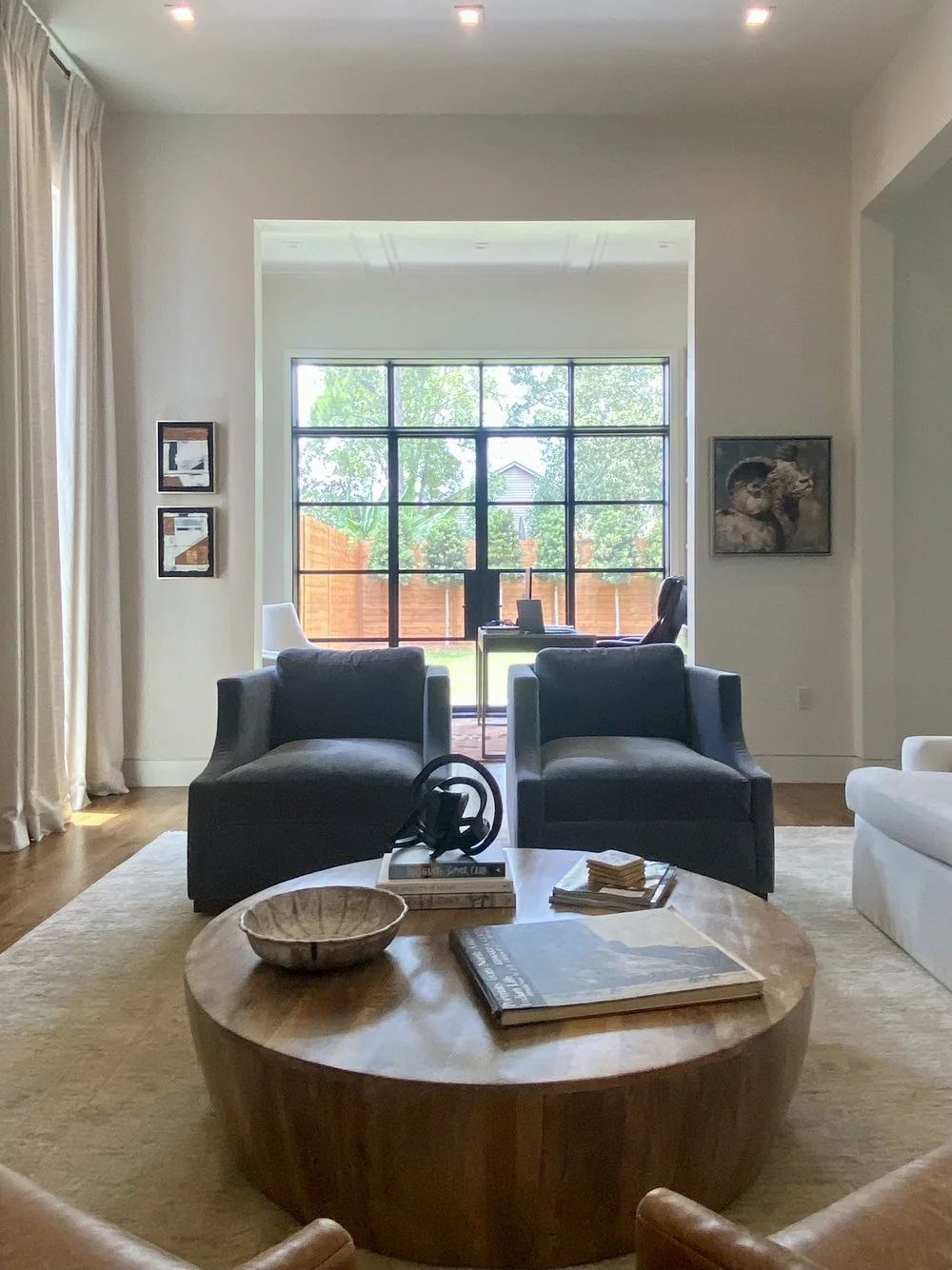 Living room with two black armchairs and a white sofa, wooden coffee table with books and a decorative bowl, large window with black frames showing a backyard with a wooden fence and trees, artwork on cream walls, beige curtains, and hardwood floor.