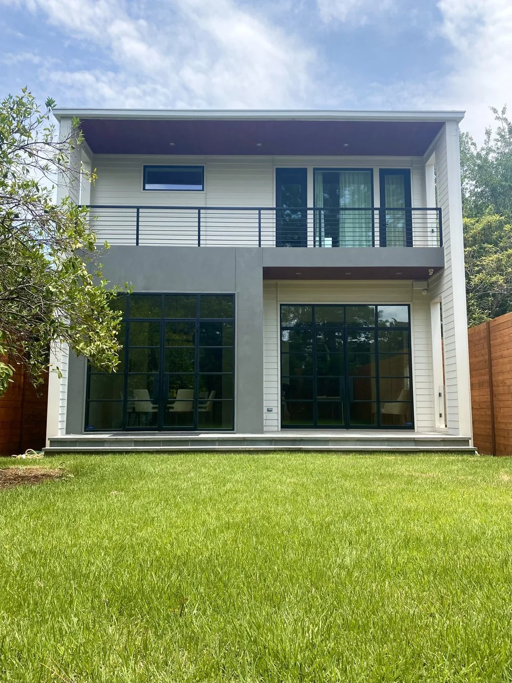Modern two-story house with large glass sliding doors, a balcony on the second floor, and a well-maintained green lawn in the backyard.