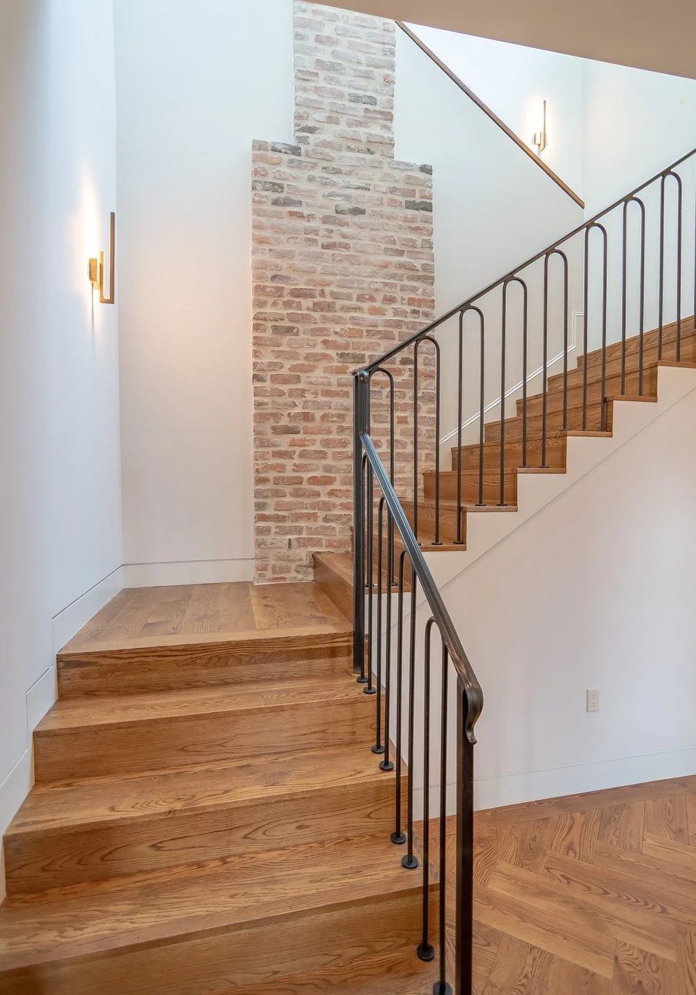 Interior staircase with wooden steps, black metal railing, white wall, exposed brick column, wall-mounted lights, and wood flooring.