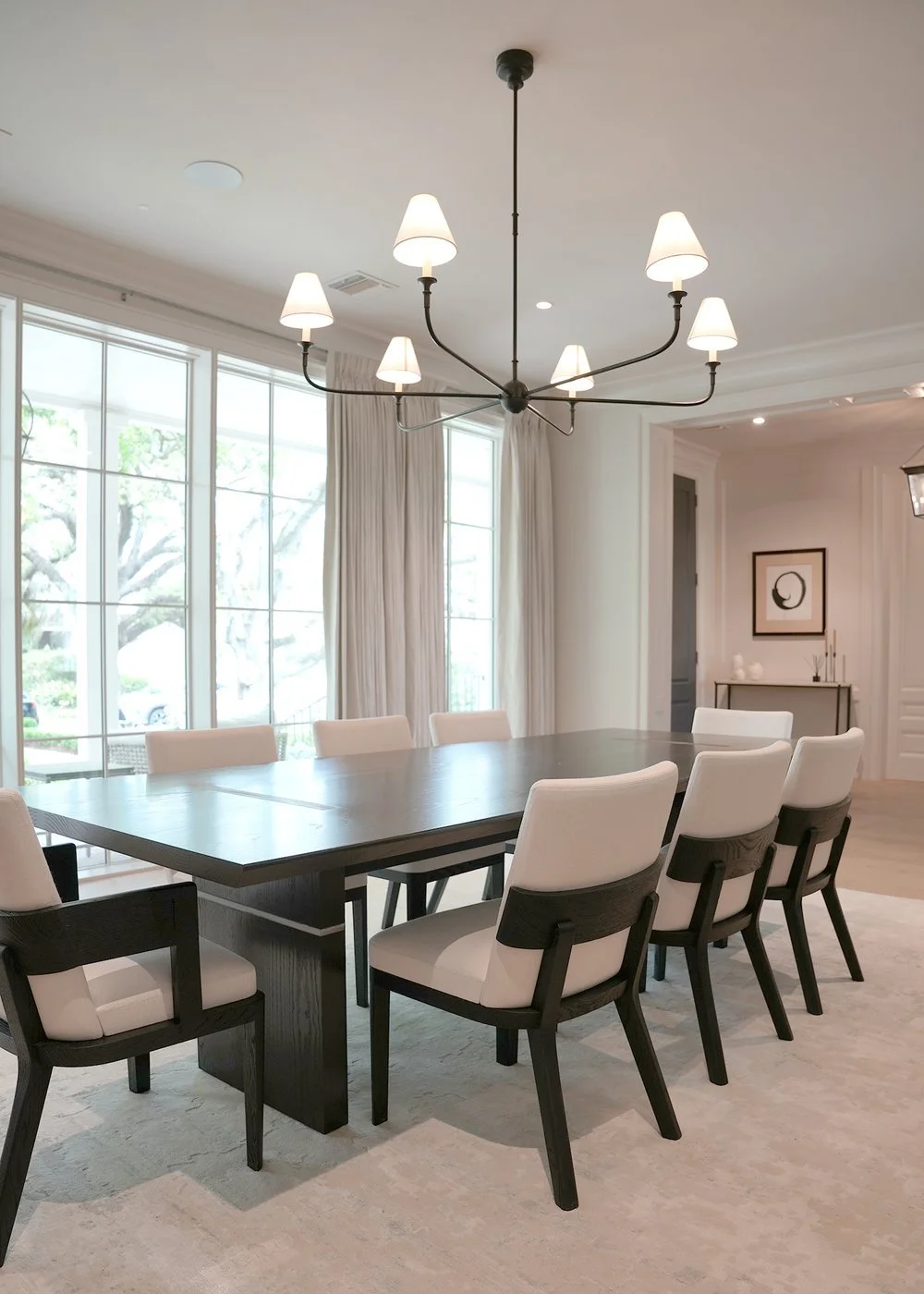 Modern dining room with a large wooden table, white upholstered chairs, large windows with curtains, and a contemporary chandelier with multiple lamp shades.