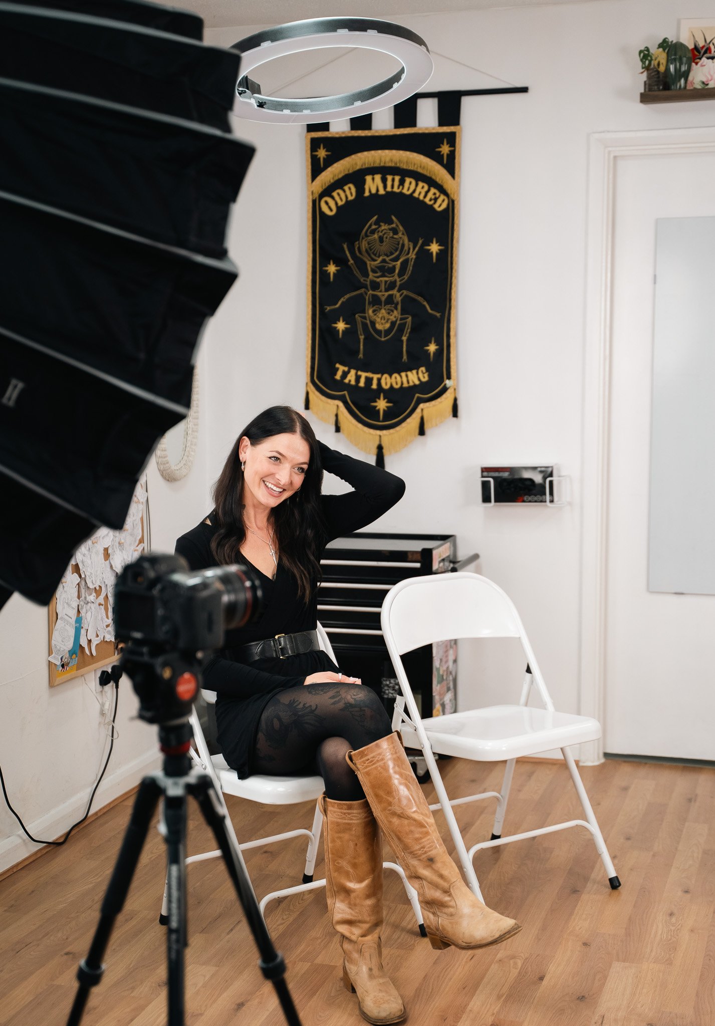 A woman sitting on a white folding chair, smiling, with her hand behind her head, in a tattoo studio. She is dressed in a black outfit and tall tan boots. There is professional camera equipment filming her. A banner with a crab and the words "Odd Mil