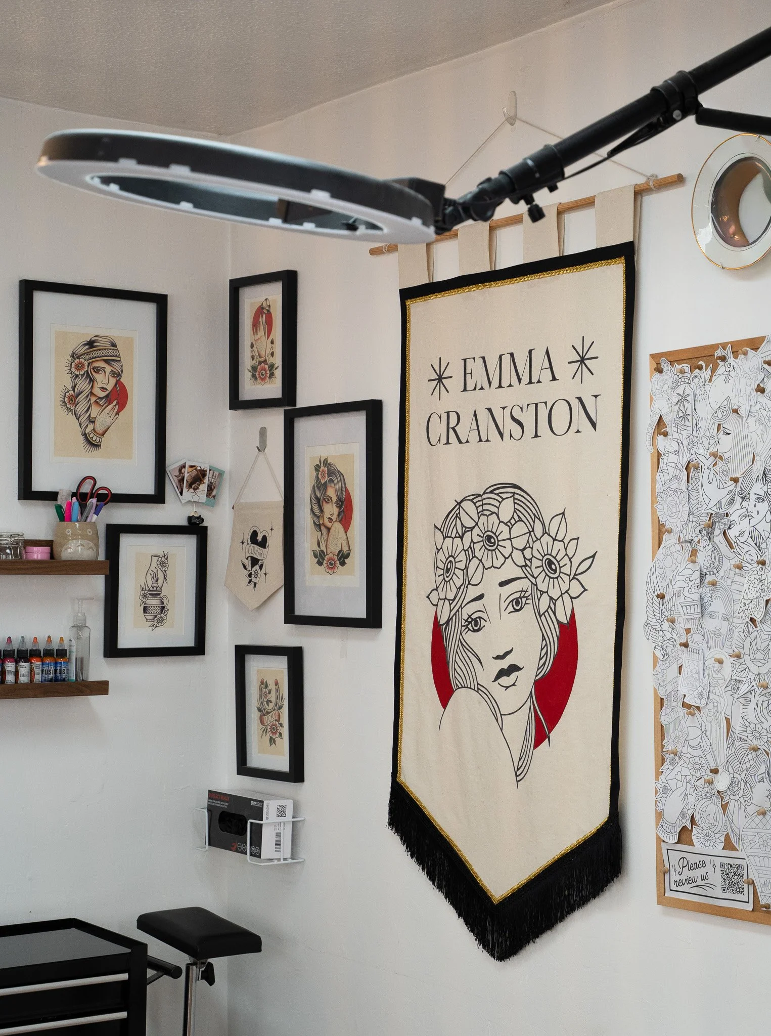 A tattoo studio space with artwork, including a large banner with the name Emma Cranston and a woman's face surrounded by flowers. The wall displays framed illustrations, a small hanging banner, spray paints, and art supplies.