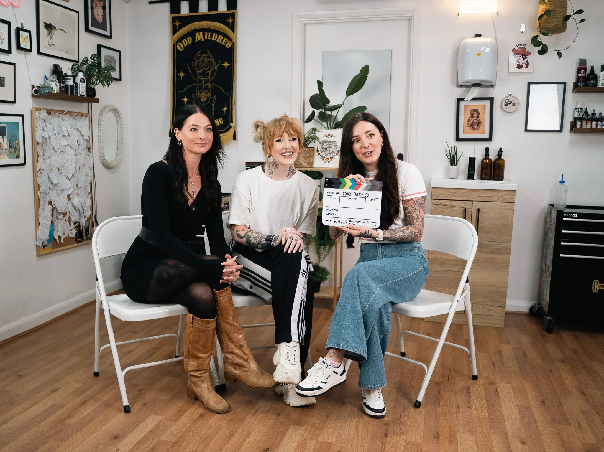 Three women sitting on white chairs in a tattoo studio, one holding a clapperboard with the words "All Your Tattoo Co." written on it, smiling and posing for the camera.