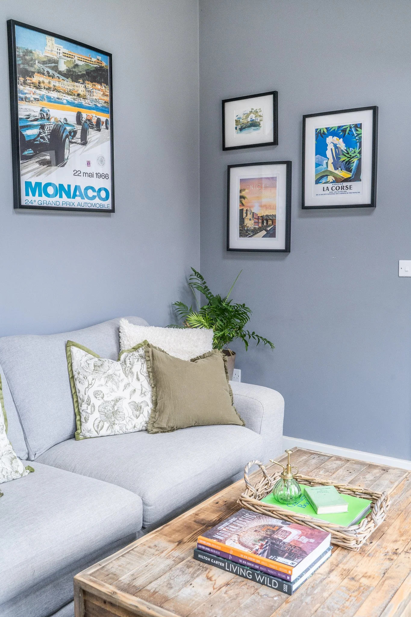 Living room with a gray sofa decorated with patterned and plain cushions, a wooden coffee table with books and a green decorative perfume bottle, and a wall adorned with five framed pictures including vintage travel posters and scenic art, with a pot