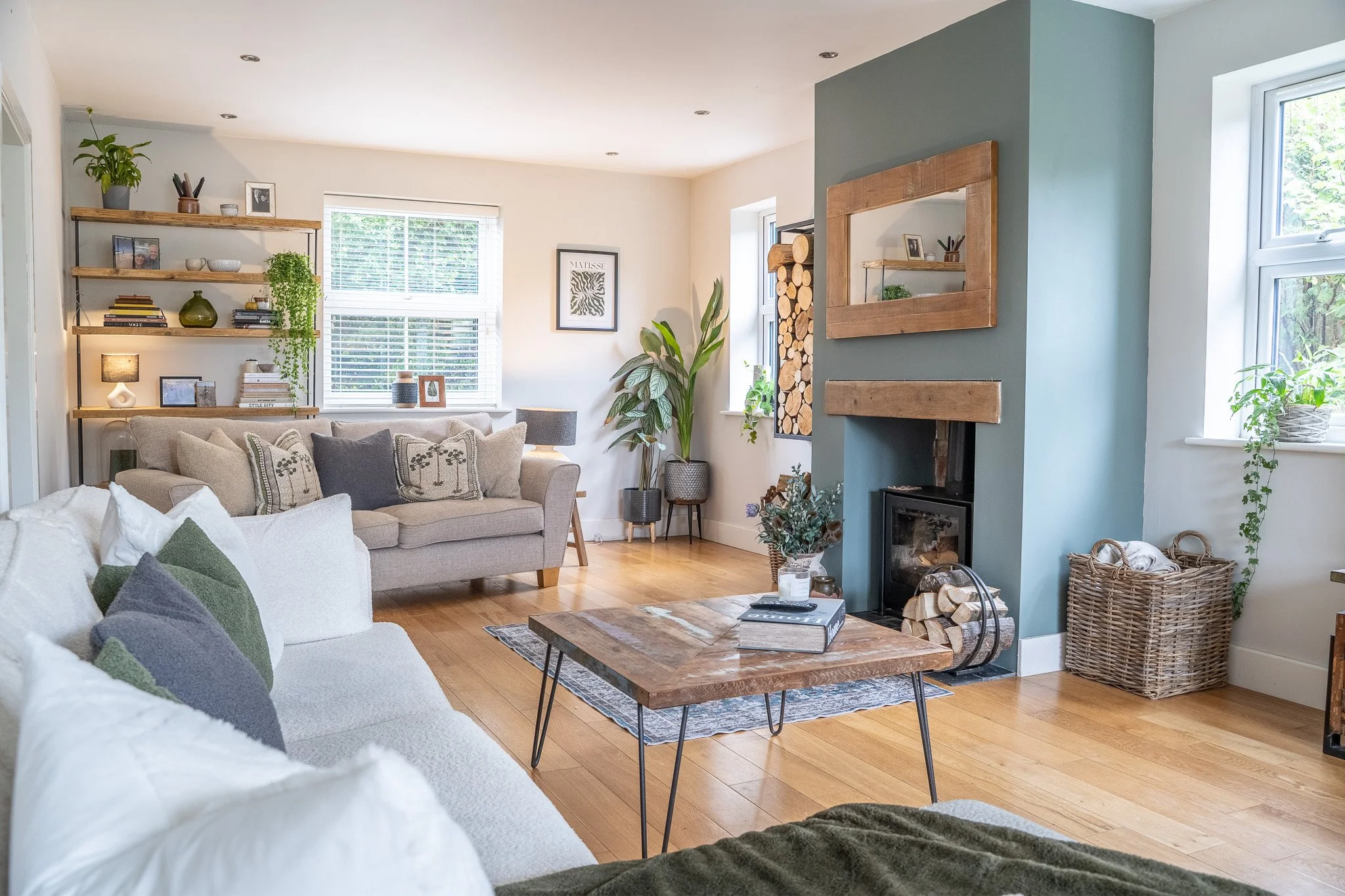 Bright living room with white and beige sofas, a wooden coffee table, a blue accent wall with a fireplace, large windows with white blinds, wooden floors, and decorative plants and shelves.