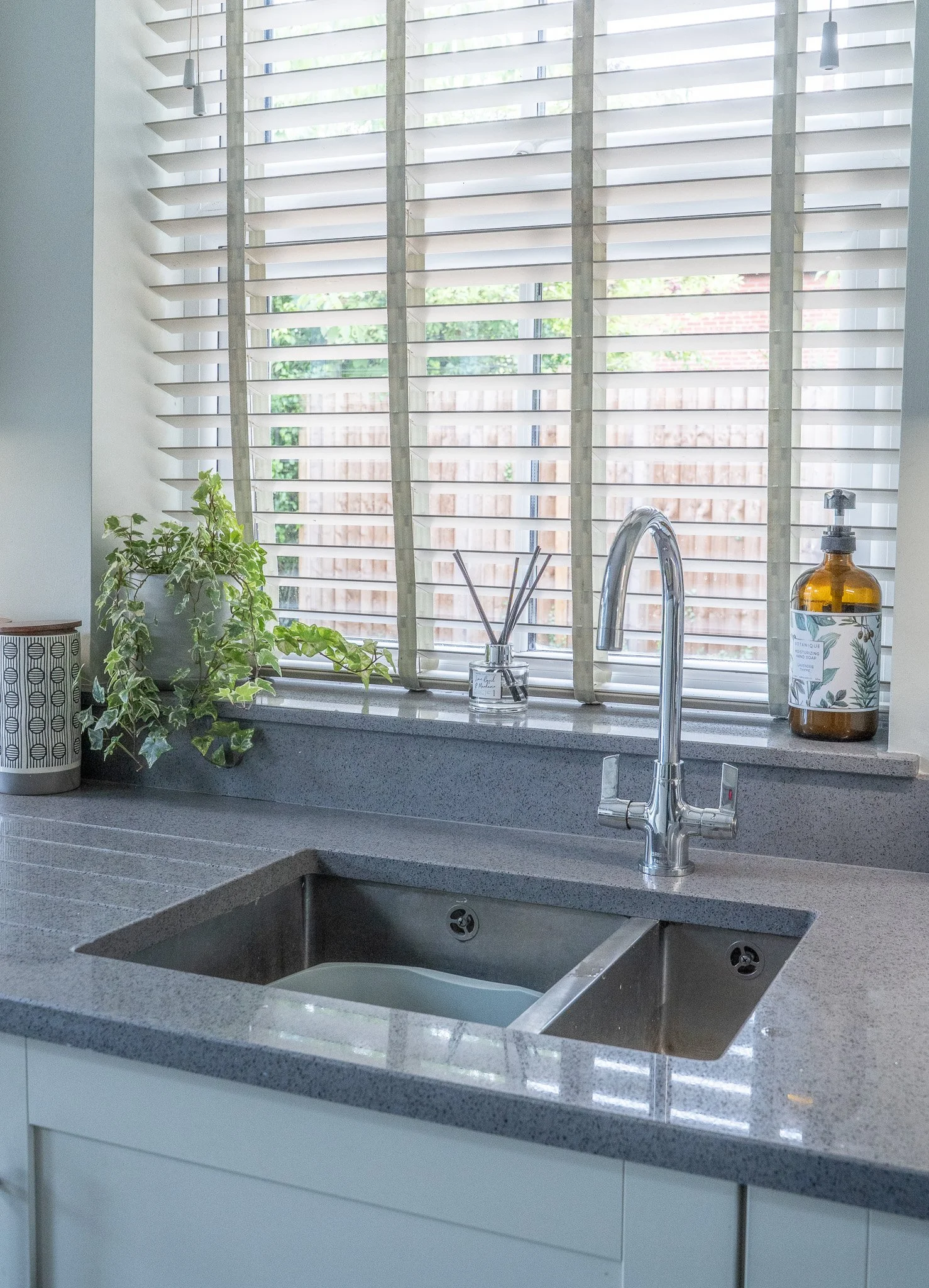 Kitchen sink with window, potted plant, diffuser, and soap dispenser.