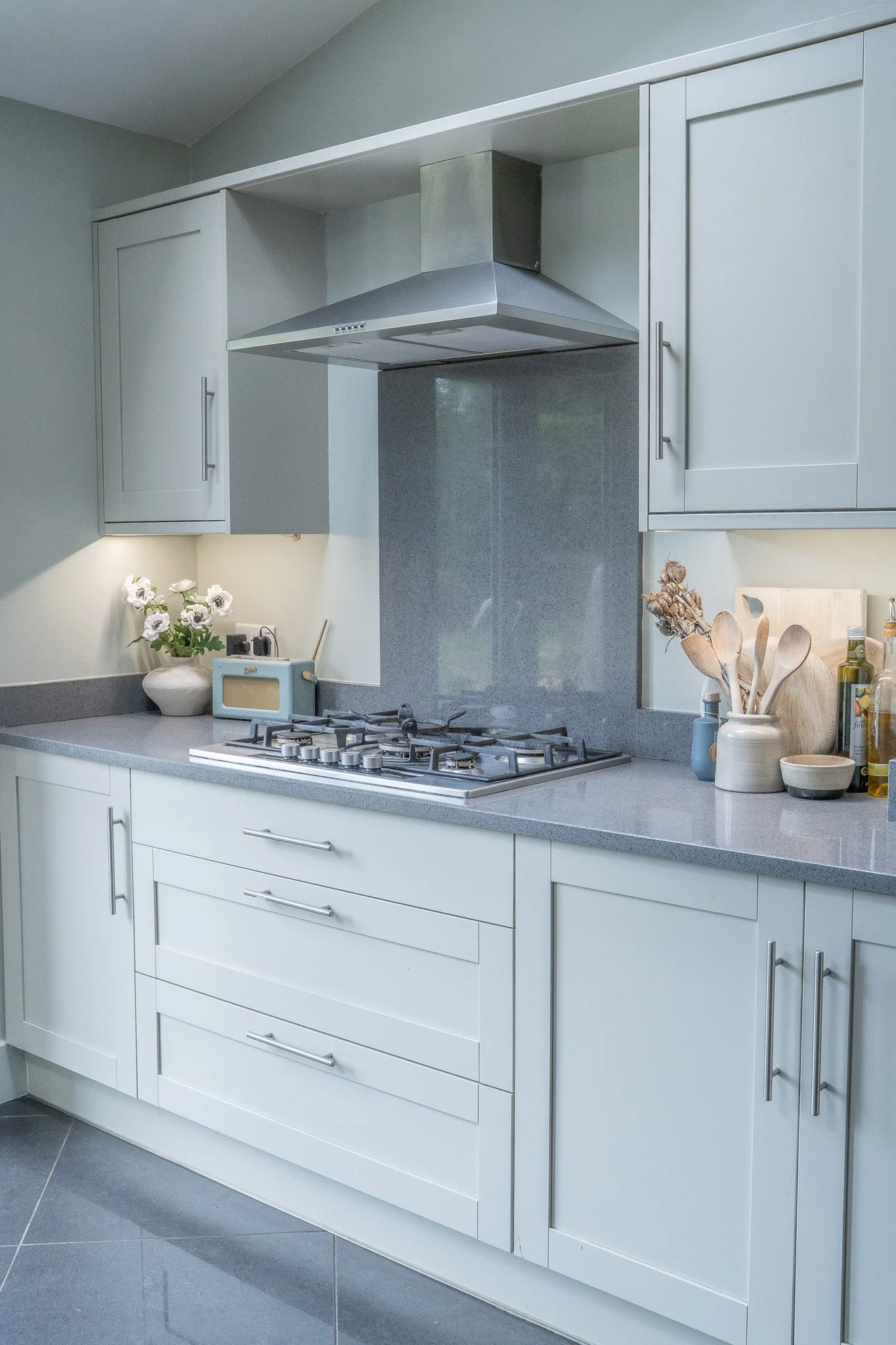 Modern kitchen with white cabinets, gray countertops, and a stainless steel range hood over a gas stove, with a vase of flowers and kitchen utensils on the counter.