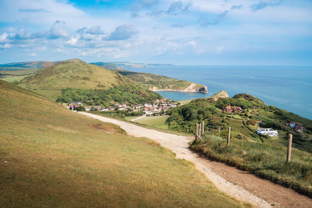 Scenic view of a coastal village with green hills, a dirt path, houses, and the ocean in the background.