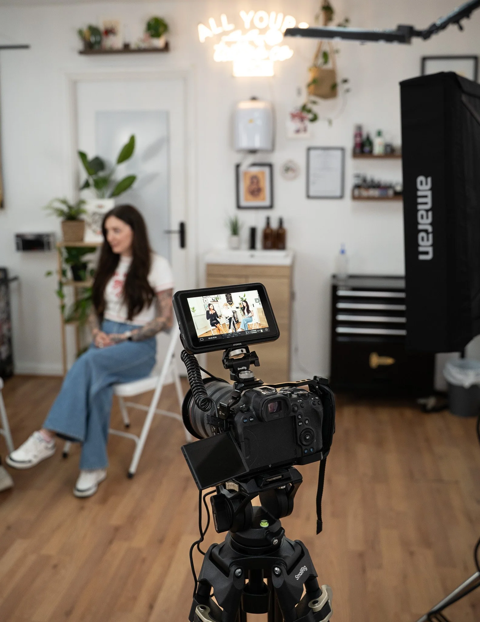 A woman with long dark hair and tattoos on her arms is sitting on a white chair in a well-lit room with wooden floors, surrounded by plants and decorative items, while being filmed by a professional camera on a tripod with a monitor showing her image