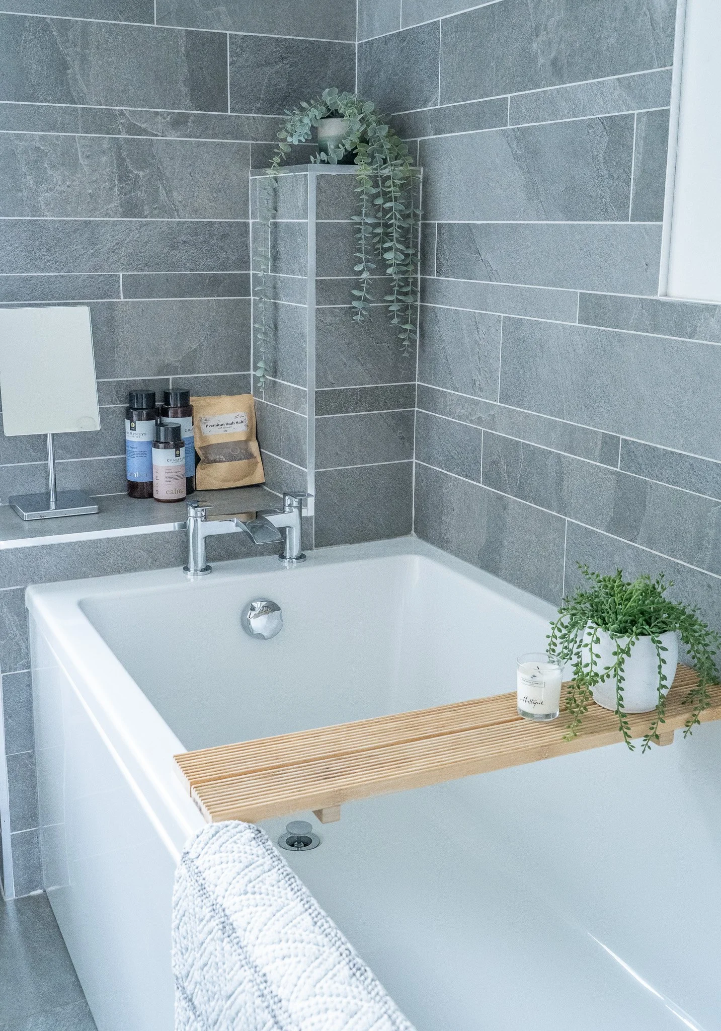A modern bathroom with a white bathtub, gray tiled walls, and a wooden bath tray holding two potted plants and a candle. There are several bottles of bath products on a small shelf, and a white towel hangs over the edge of the tub.