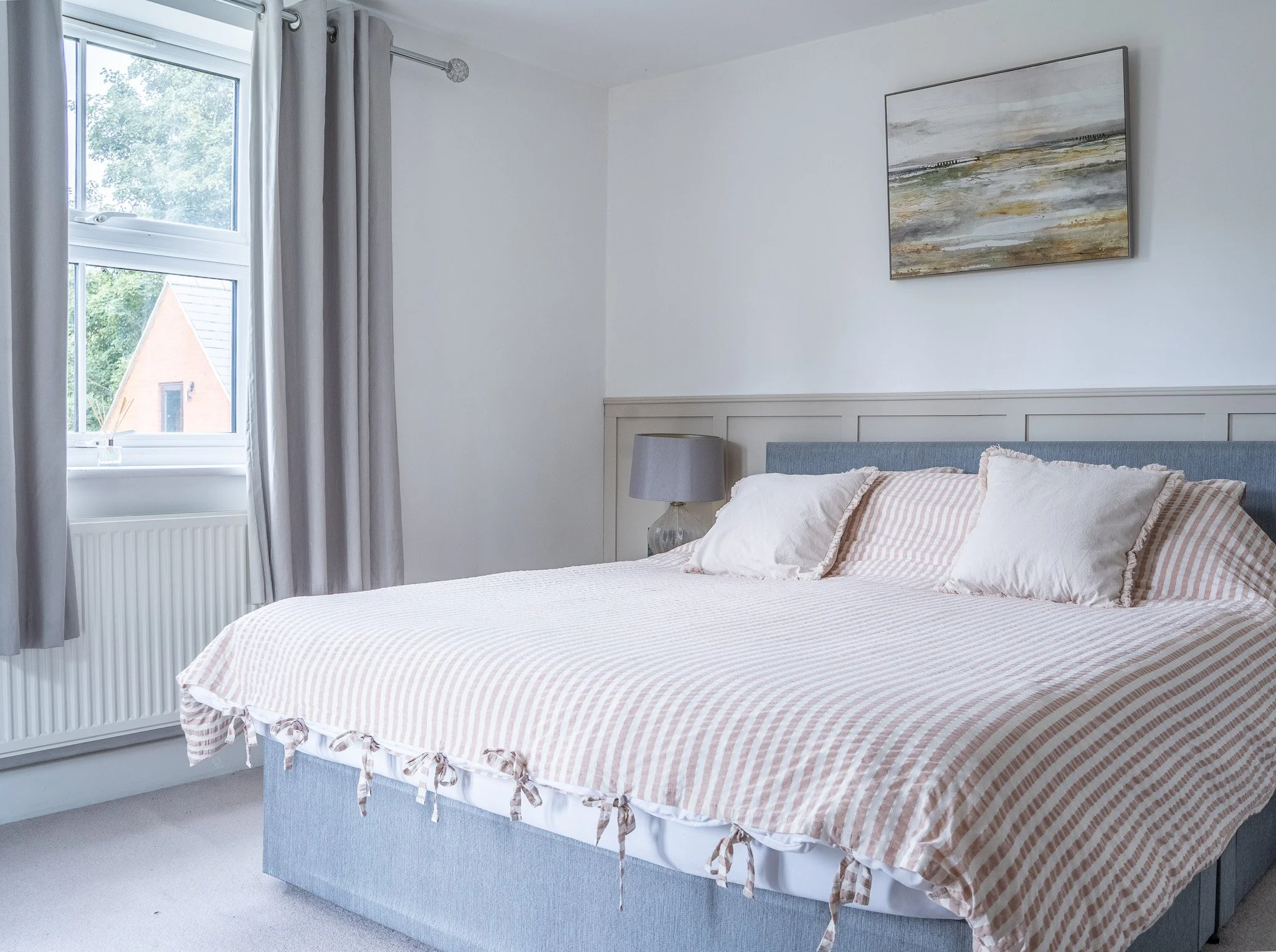 A bedroom with a bed that has a beige and white striped duvet and pillows, a gray headboard, a gray nightstand with a gray lamp, a window with gray curtains, and a landscape painting on the wall.