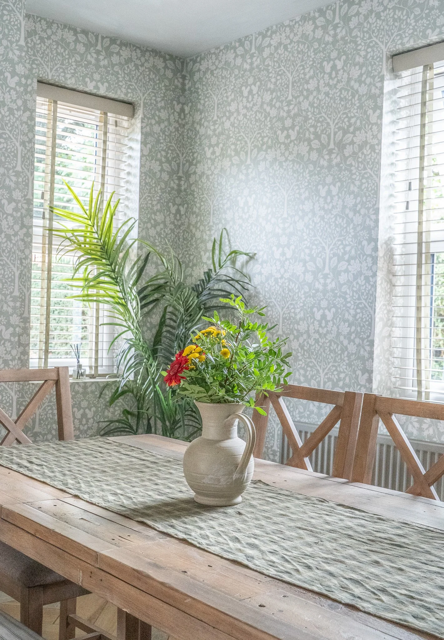 A dining room with a wooden table, floral table runner, ceramic pitcher with colorful flowers, green houseplants, patterned wallpaper, and windows with blinds letting in natural light.