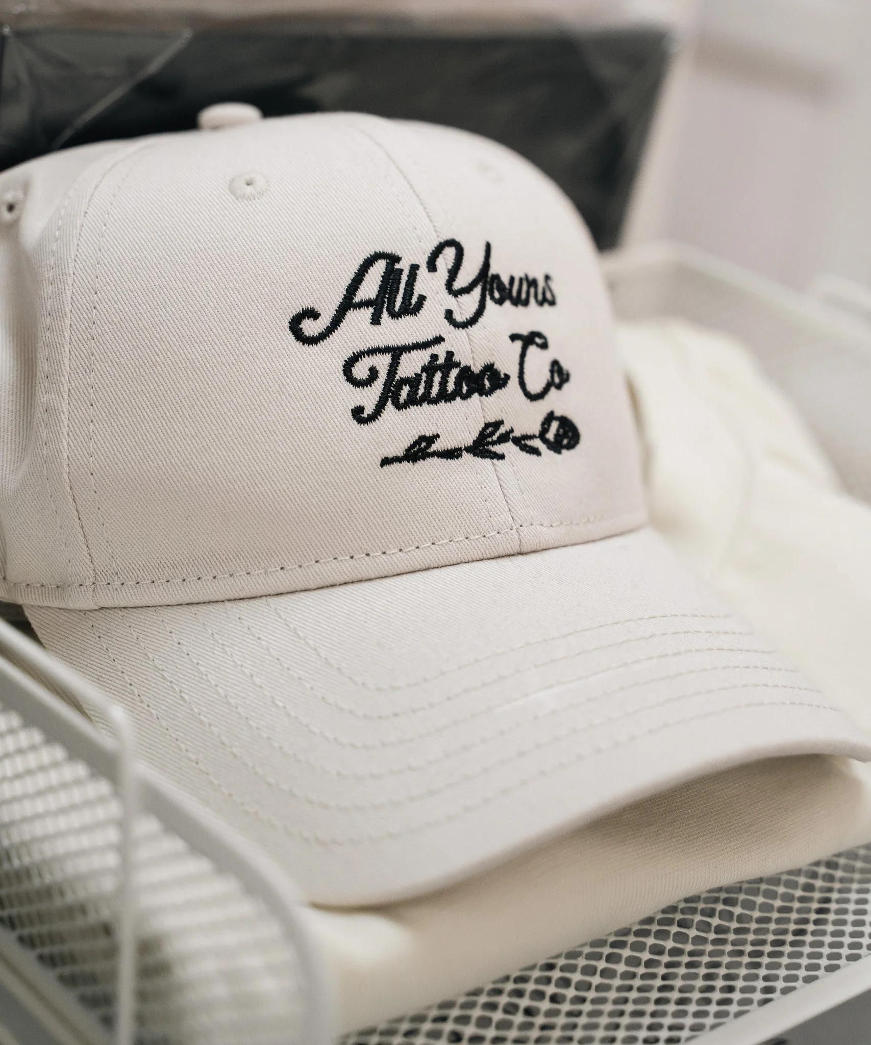 White baseball cap with black embroidery that reads 'All Yours Tattoo Co' in cursive font.