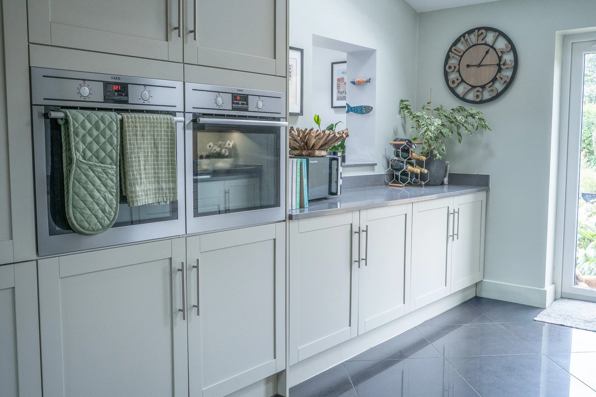 Modern kitchen with white cabinets, gray countertops, built-in oven, oven mitts hanging, potted plant, wine rack, wall clock, and sliding glass door.