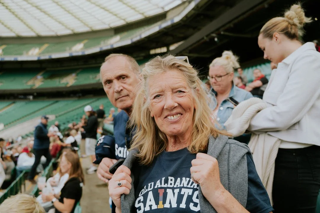 A smiling older woman wearing a 'Old Albany Saints' t-shirt at a sporting event, surrounded by other spectators in a stadium.