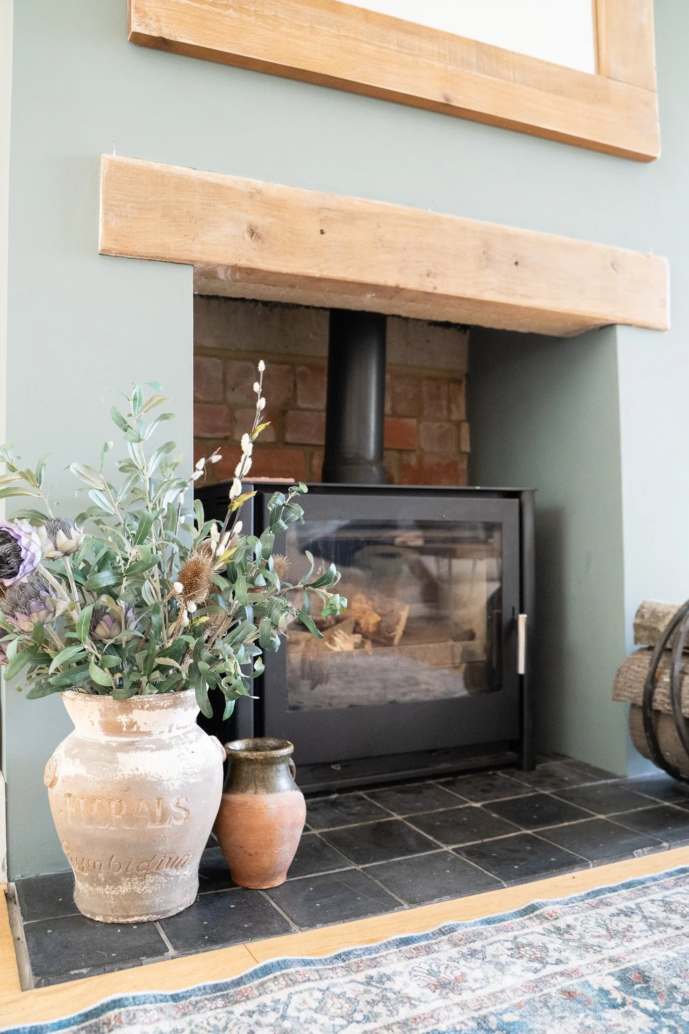 A cozy room featuring a fireplace with a wood-burning stove, a wooden mantel, and a brick backdrop. A large decorative vase with lavender and greenery, alongside a smaller pottery vase, are placed on the dark tiled hearth. Part of a patterned rug and