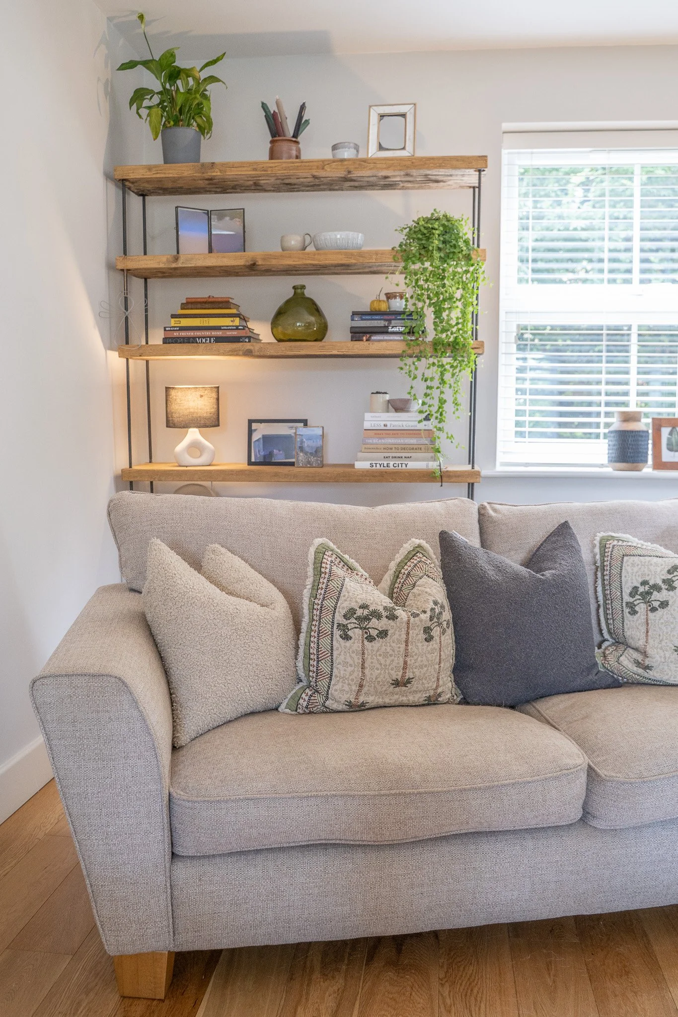 Living room with a cozy beige sofa adorned with decorative pillows, a wall-mounted wooden shelf displaying books, plants, and decorative items, a window with blinds allowing natural light, and a small table lamp on a side table.