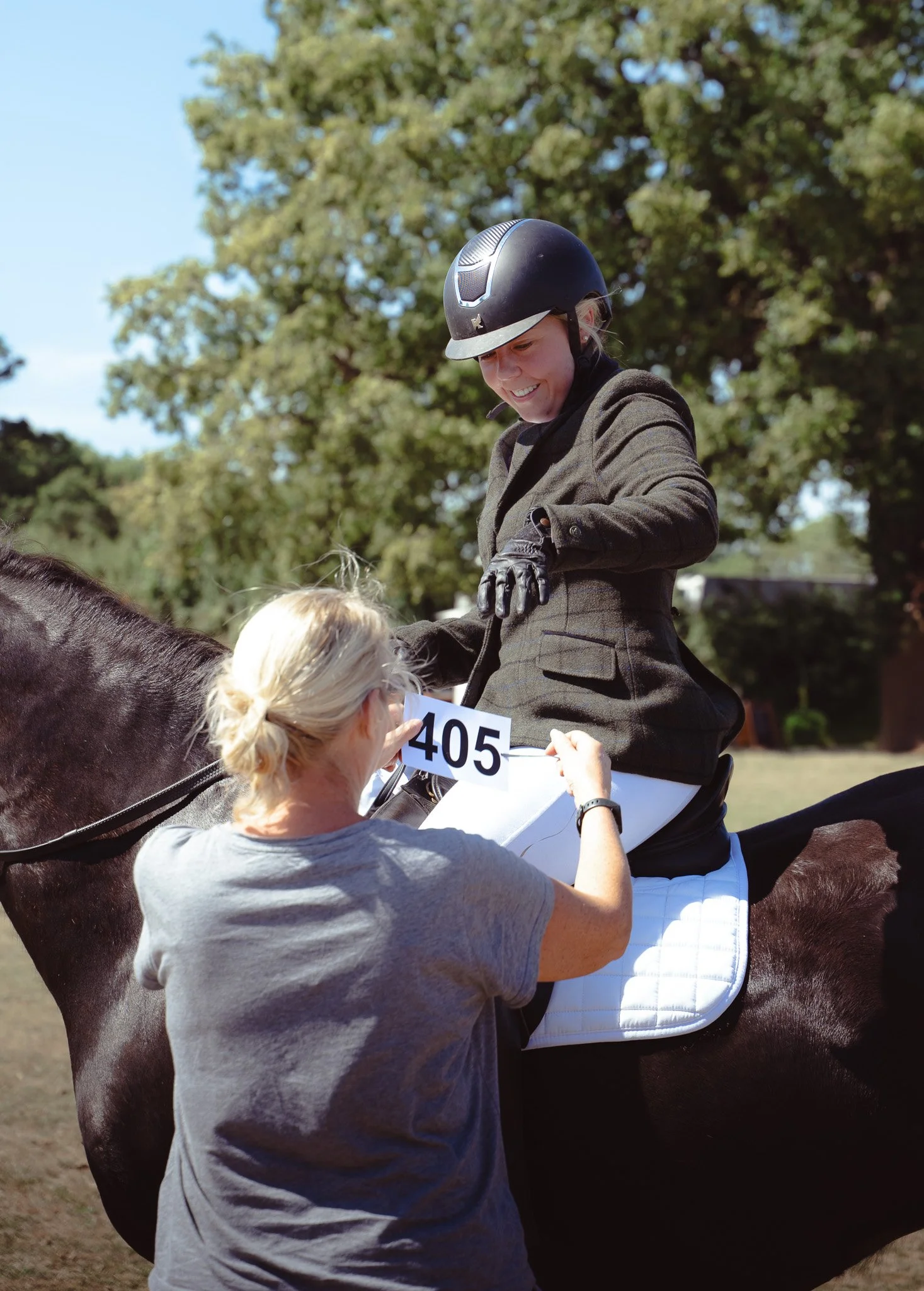 A woman in a competition helmet and formal riding jacket smiling on a dark horse, with a person holding a sign with the number 405, in an outdoor setting with trees in the background.